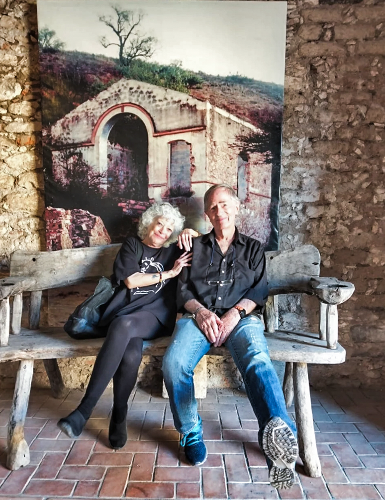 An elderly woman and man sitting on a rustic wooden bench with armrests, smiling and posing together. Behind them is a large photograph of an old stone building with an arched entrance, set in a natural landscape with trees and hills.