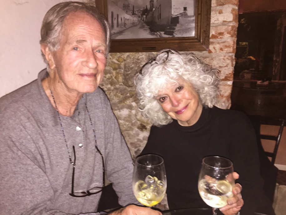 An elderly man and a woman with curly gray hair sitting at a table, smiling, each holding a glass of white wine with lemon slices, in a restaurant with brick wall and framed picture in the background.