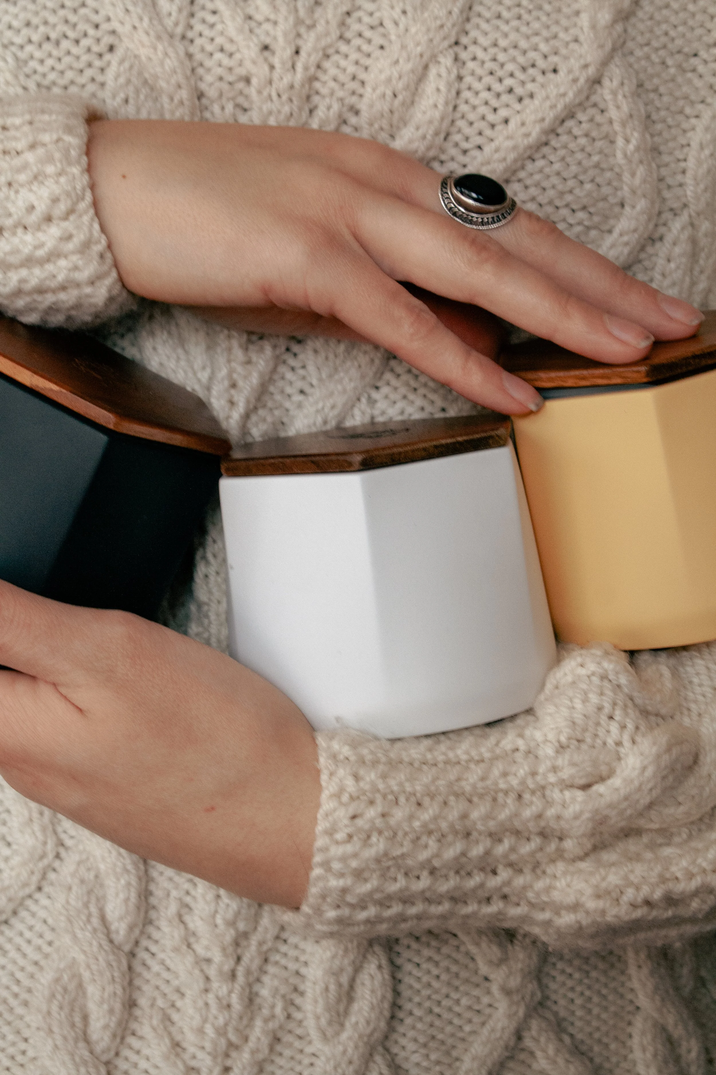Hand with gemstone ring and sweater holding three Malua ceramic cannabis storage jars in white, yellow, and black — featuring wooden lids, split compartments, and airtight seals for stylish, organized storage.