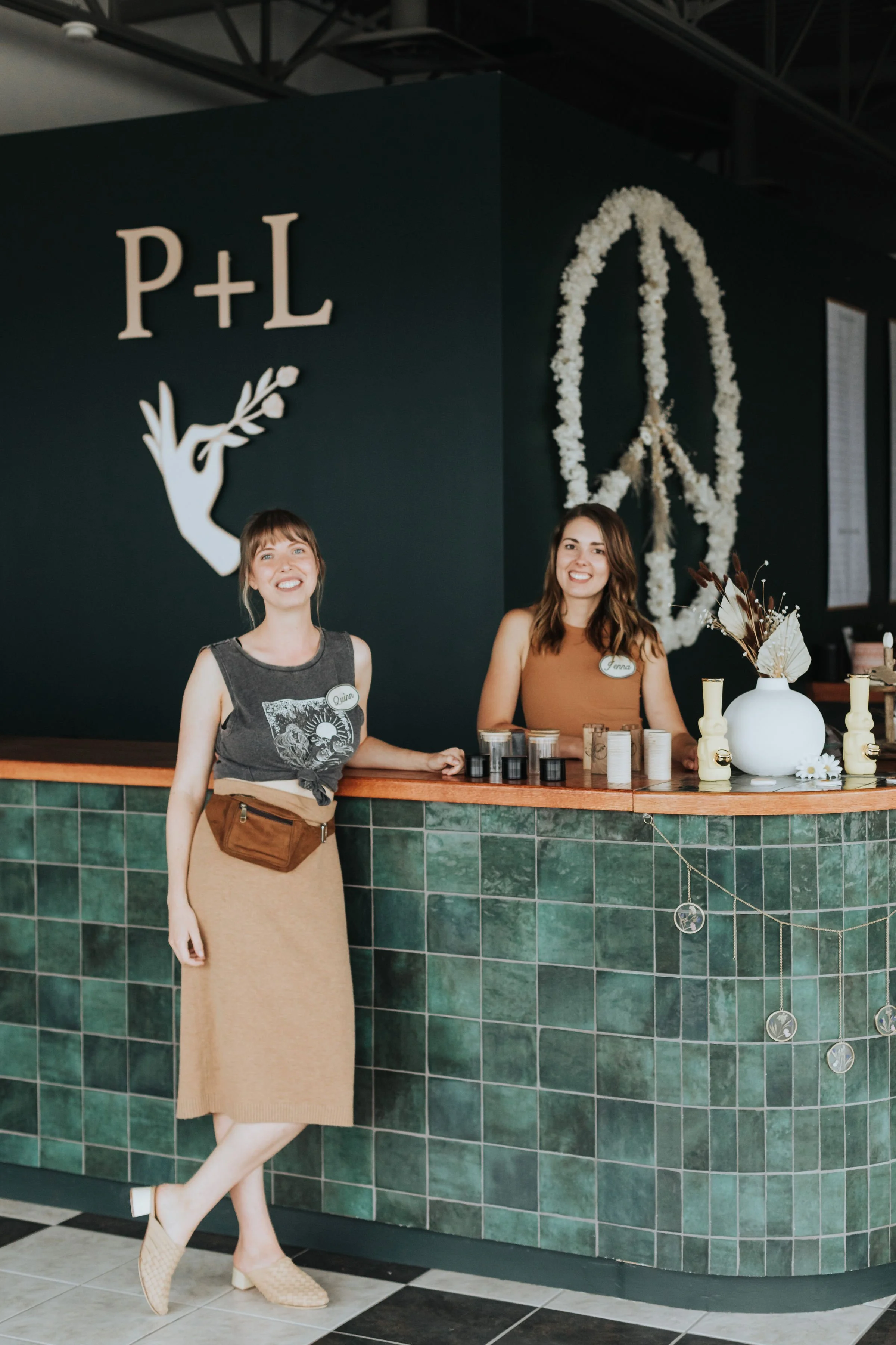 Prairie and Luna owners Quinn and Jenna behind the cash desk, surrounded by deep green tiles, soft lighting, and a dried floral peace sign in our welcoming storefront.