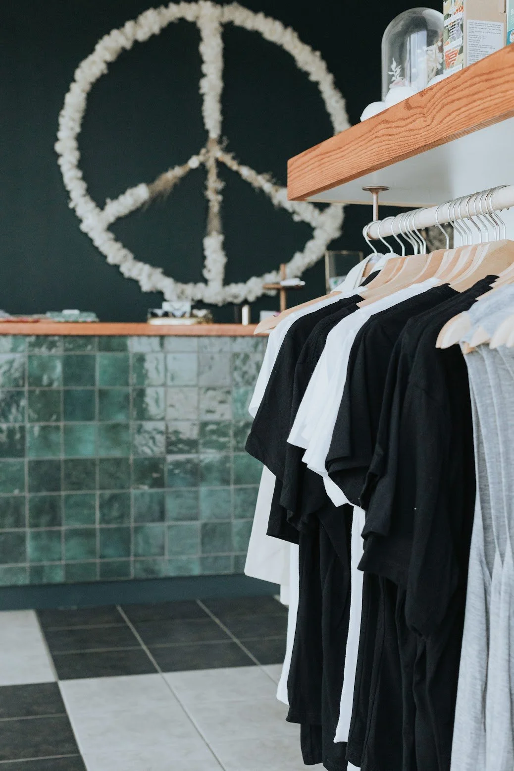 Prairie and Luna merch display featuring neutral-toned tees on a rack, set against deep green walls and a dried floral peace sign by a local artist.