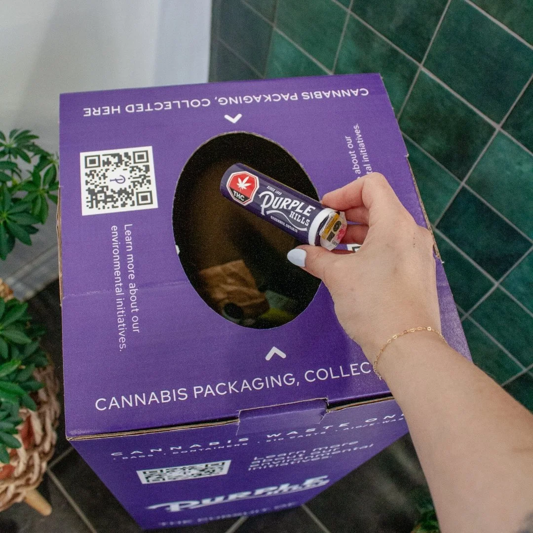 Picture of a woman's hand holding a plastic, tubular package with the Purple Hills logo on it. She is holding it over a purple recycling bin, which reads "cannabis packaging, collected here"