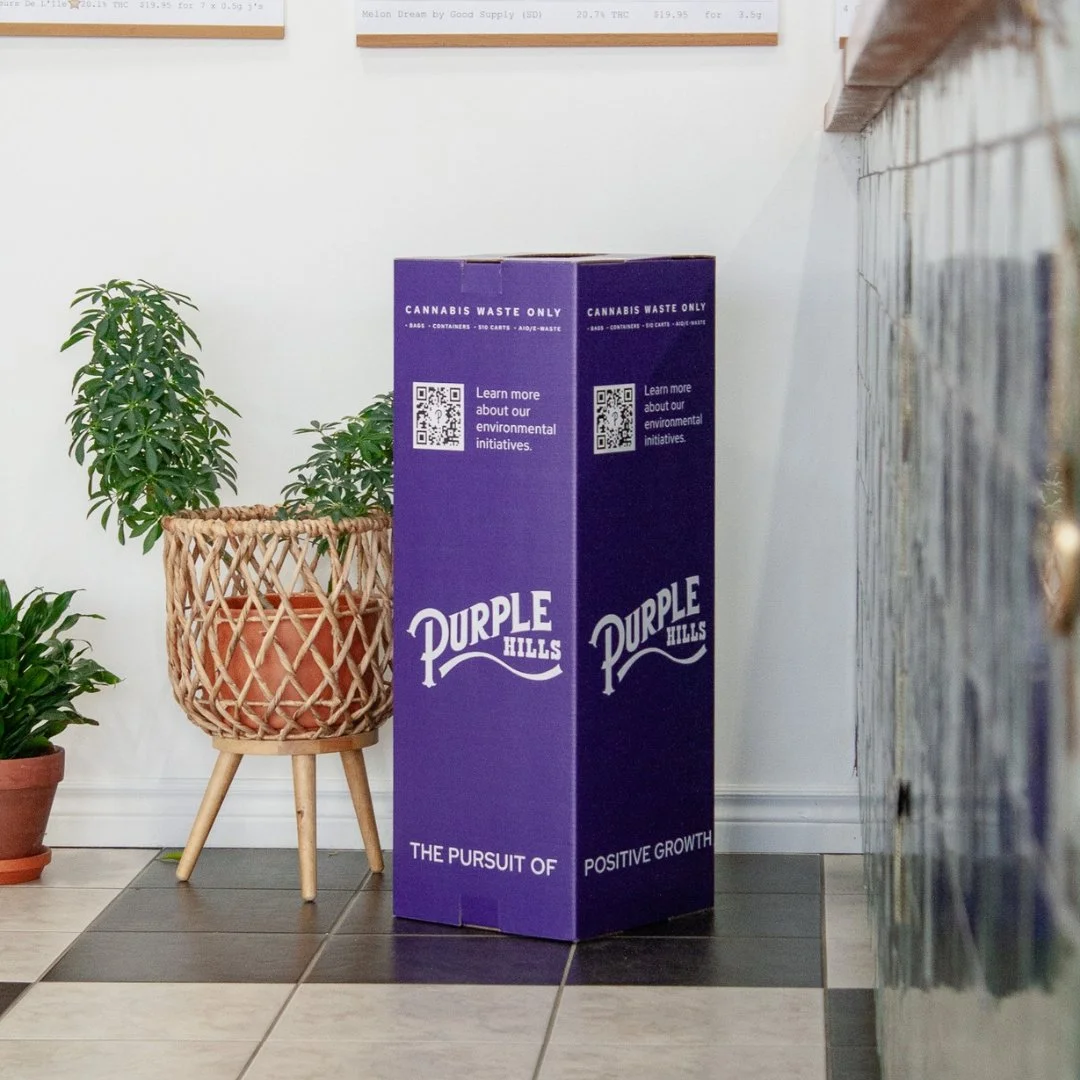 A photo of a tall, purple cardboard recycling box, with Purple Hill's logo on it. The bin is surrounded by plants and reads "cannabis waste only."