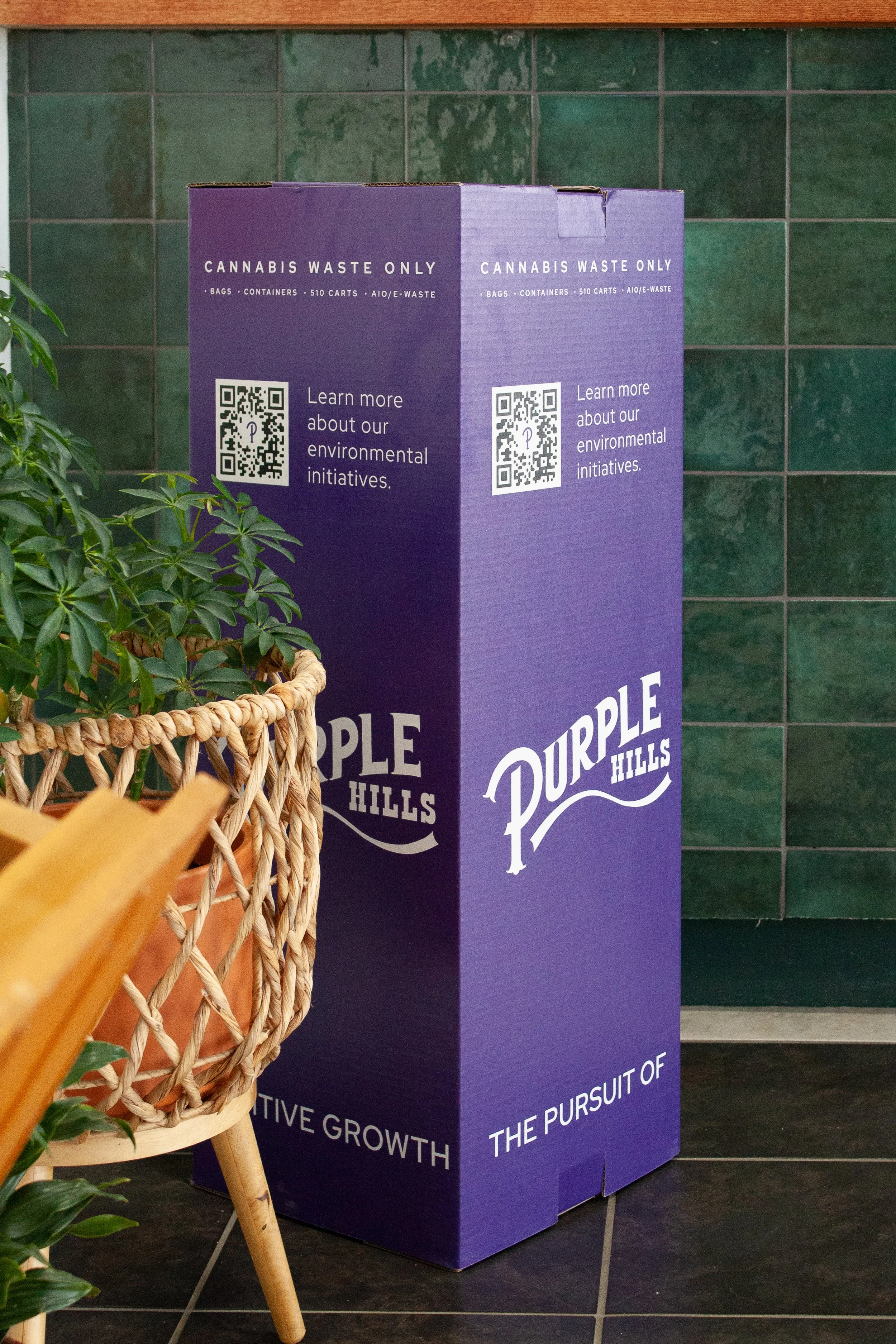 Large purple box labeled 'CANNABIS WASTE ONLY' with QR code and environmental initiative information, next to potted plant and green tiled wall.