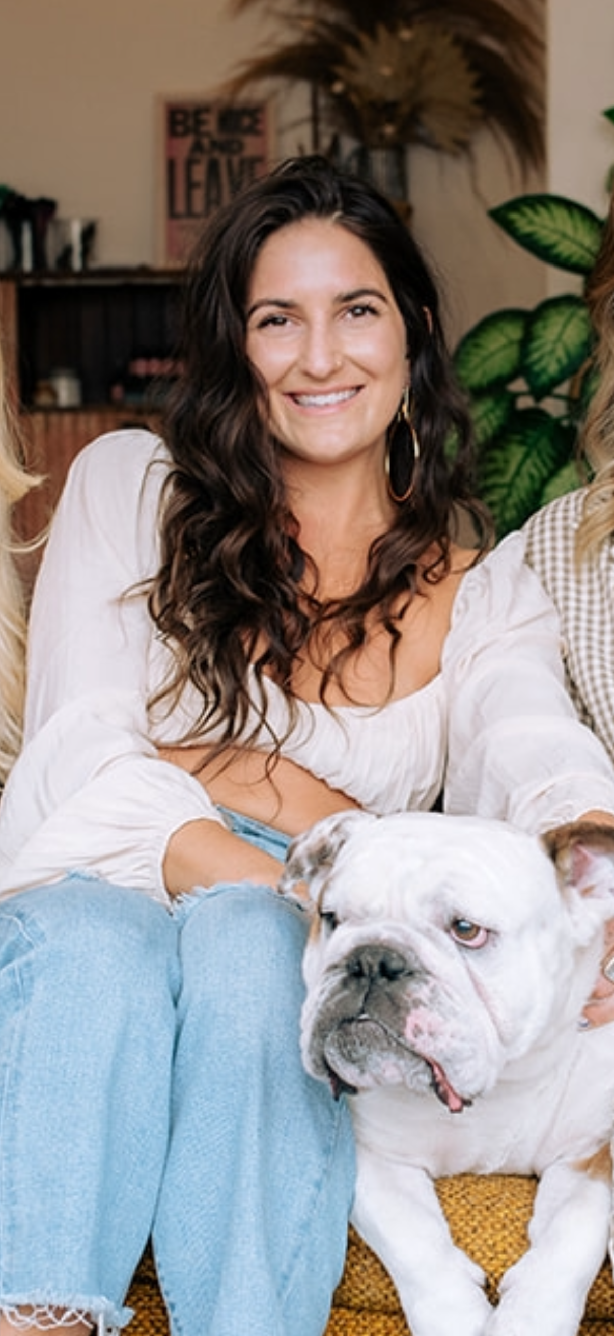 A woman with long, dark, curly hair smiling while standing indoors, wearing a white crop top and high-waisted jeans, with plants and furniture in the background.