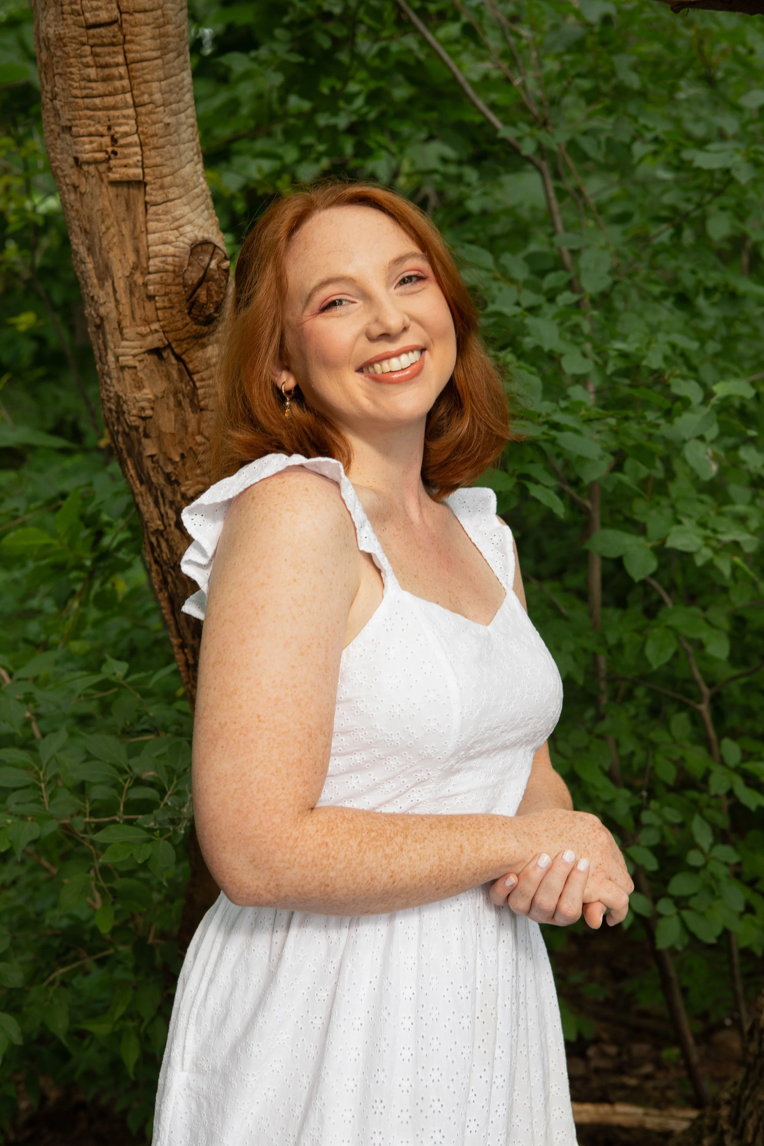 Red headed smiling woman in white sundress in front of tree trunk and green ivy.