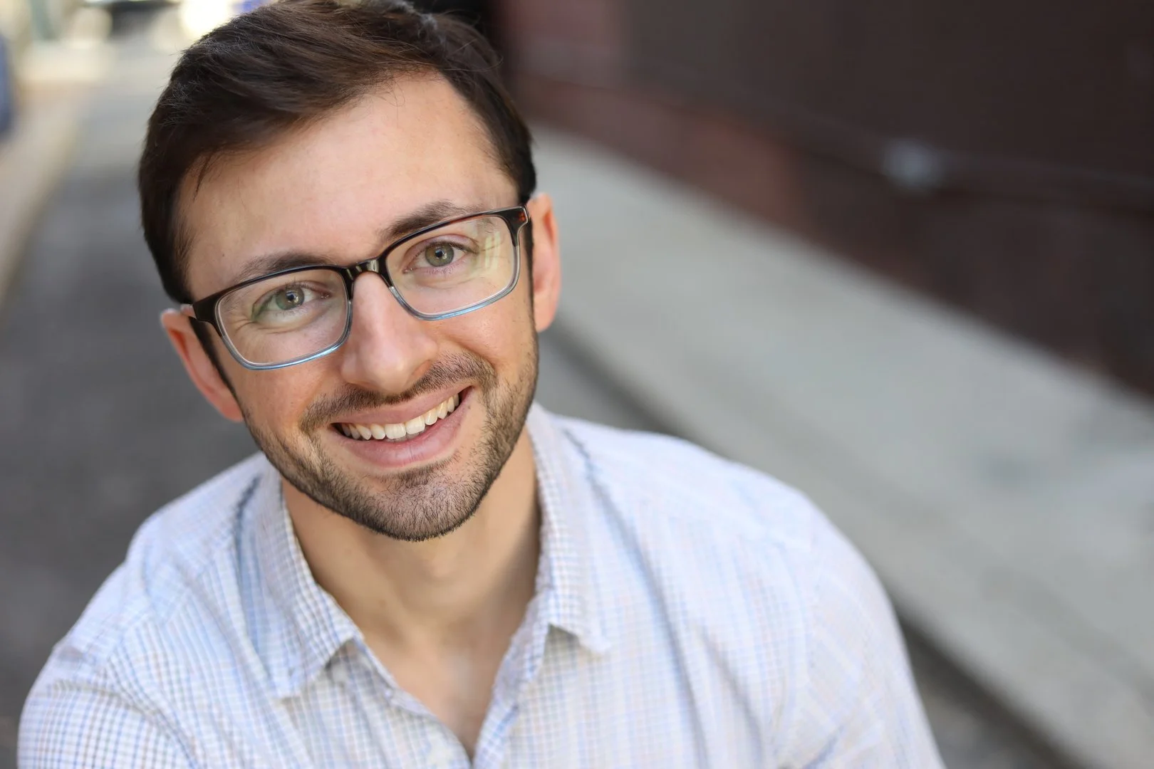 Smiling man with glasses, mustache and beard standing in front of blurred sidewalk.