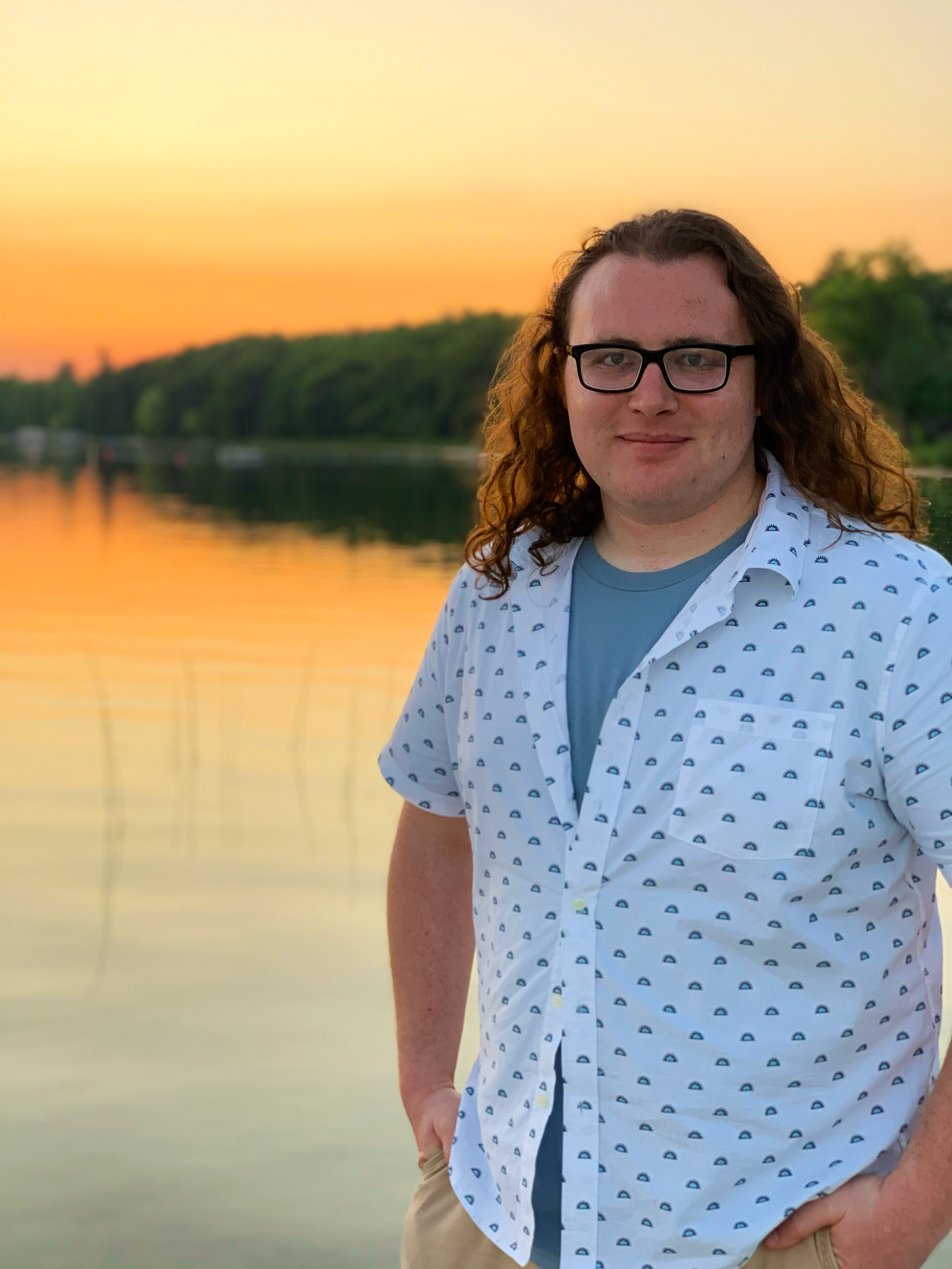 Young man with long red hair and glasses standing in front of evening pond view with sunset in background