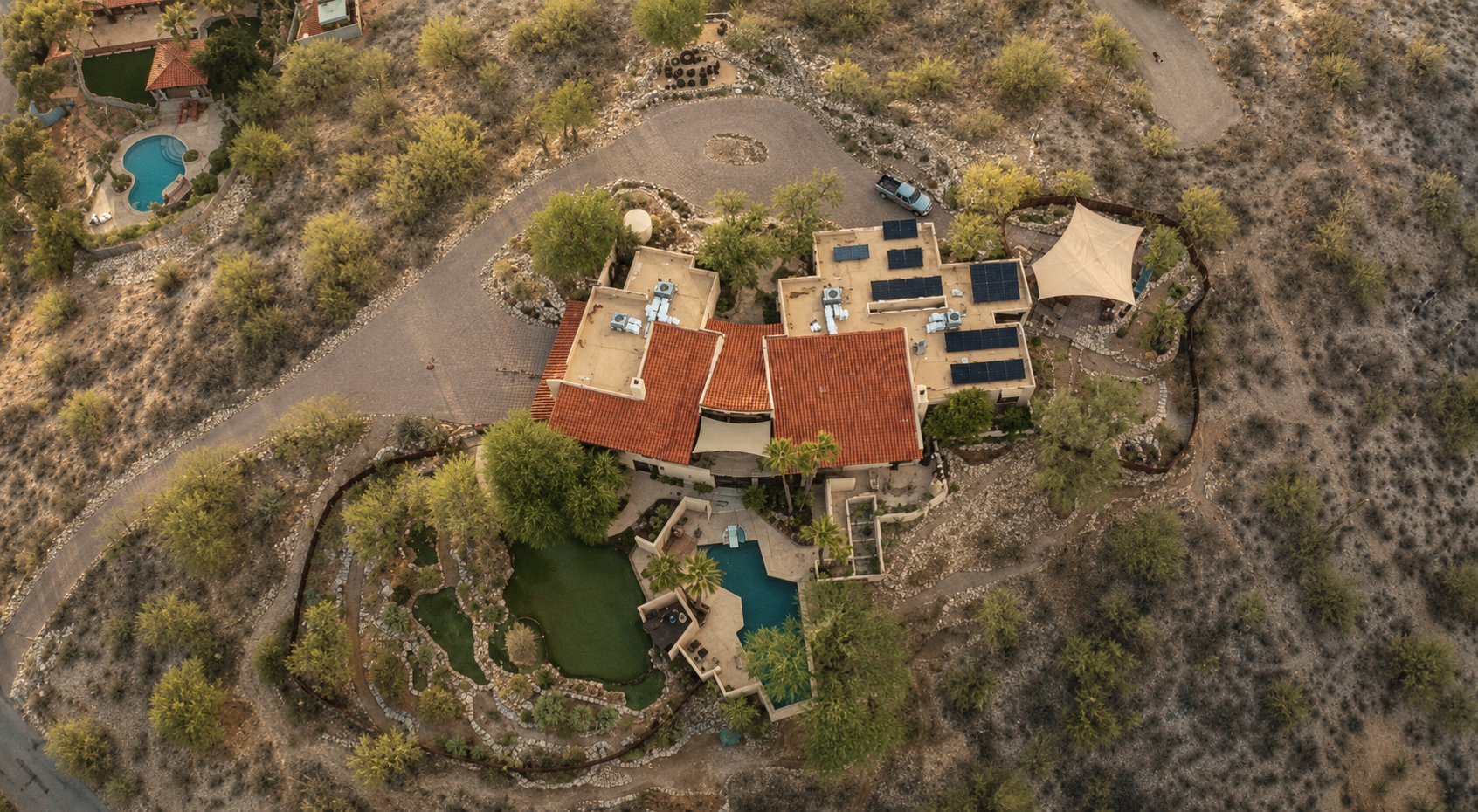 An aerial view of a large house with a red-tiled roof, a swimming pool, sun loungers, and surrounding trees in a desert landscape with sparse vegetation, dirt pathways, and neighboring homes with pools.