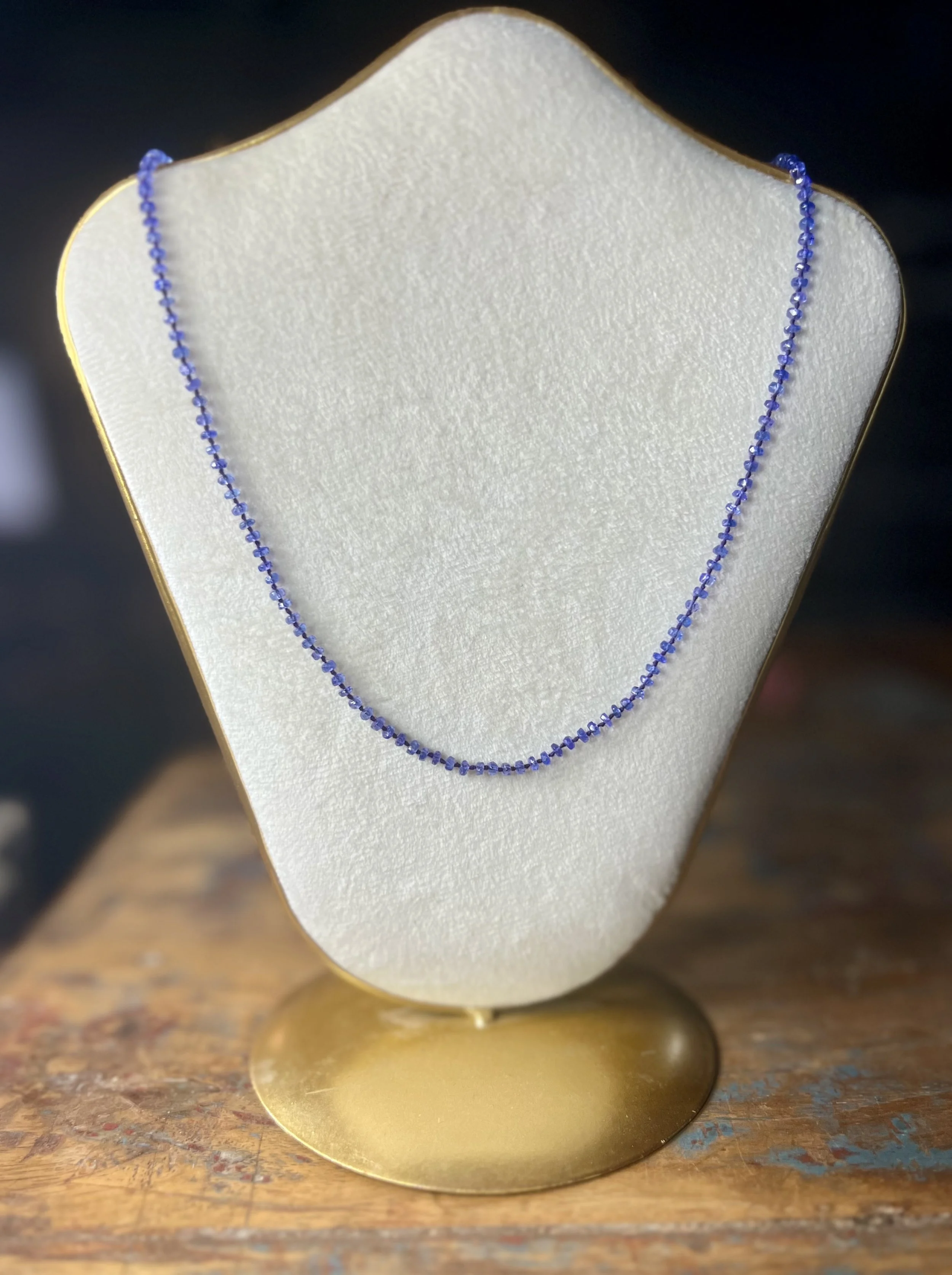 A blue sapphire necklace displayed on a white jewelry bust with a gold trim, placed on a wooden surface.