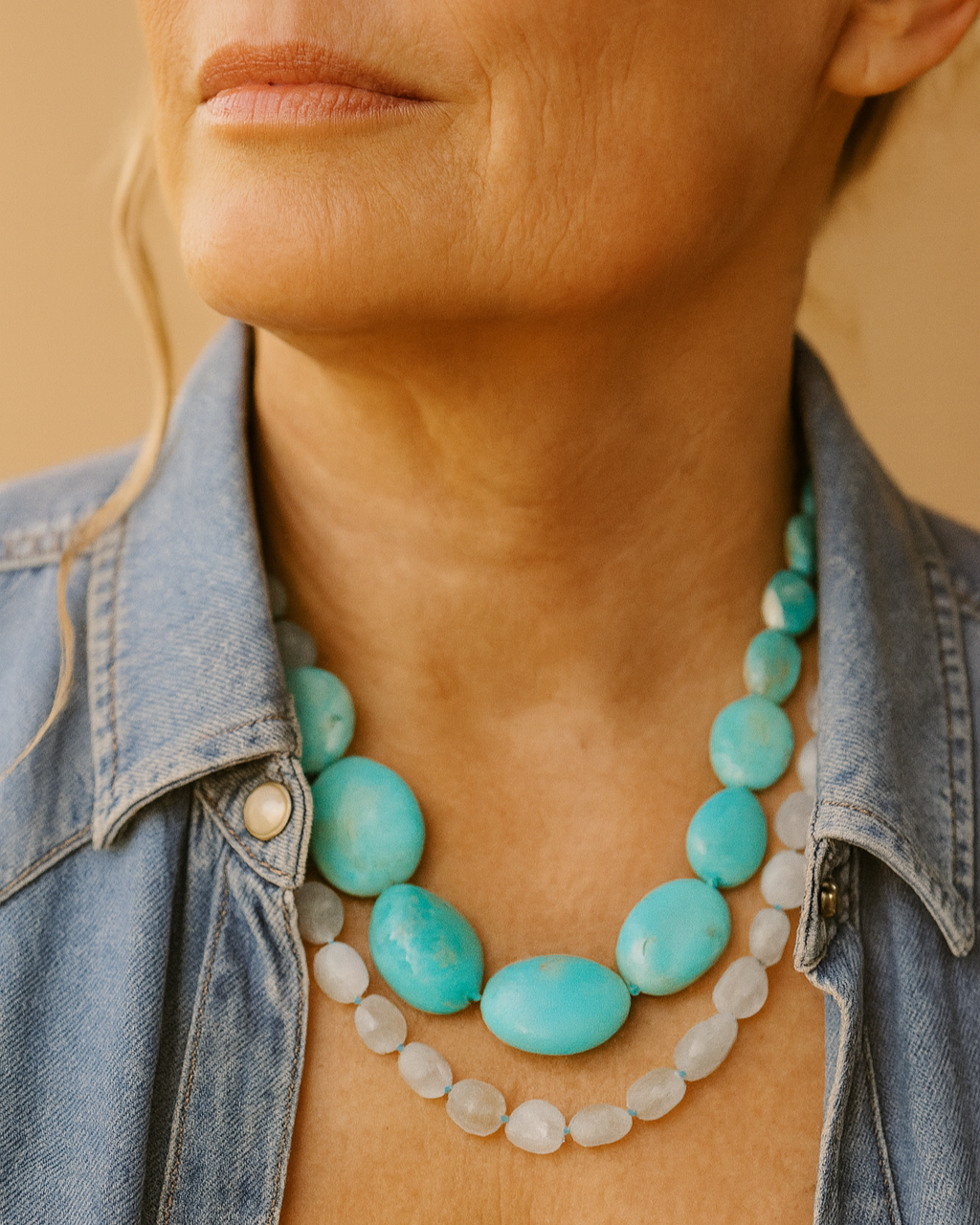 Close-up of a woman’s neck and lower face, wearing multiple turquoise and white beaded necklaces, and a denim shirt.