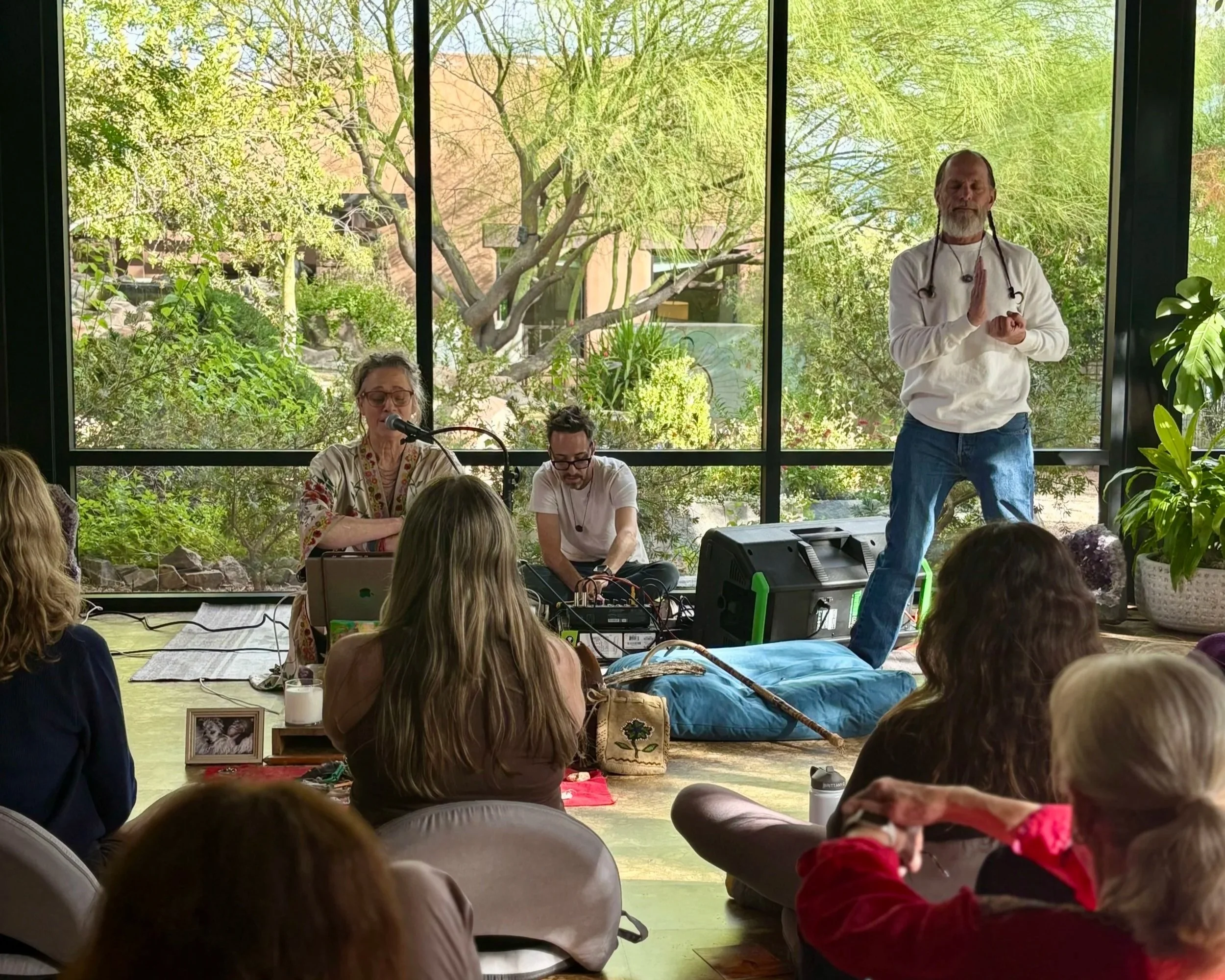 A group of people gathered in a room with large windows and surrounded by plants, watching a man standing and meditating while wearing earphones. There are musicians with equipment in the background.