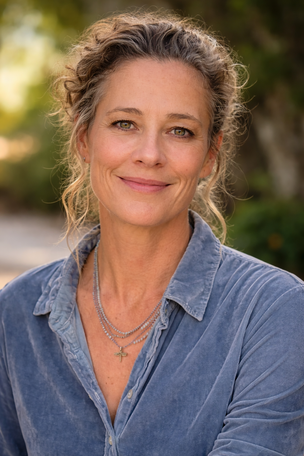 A smiling woman with light brown, curly hair, wearing a blue denim shirt and layered jewelry including necklaces with a small cross pendant, outdoor setting with greenery and blurred background.