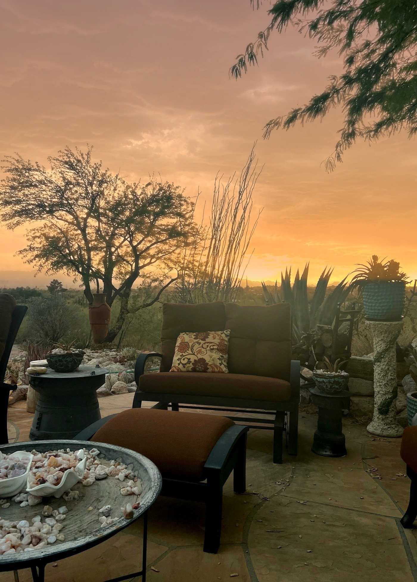 A sunset view over a patio with outdoor furniture, trees, and plants, including a table with shells, a chair with cushions, and decorative urns.