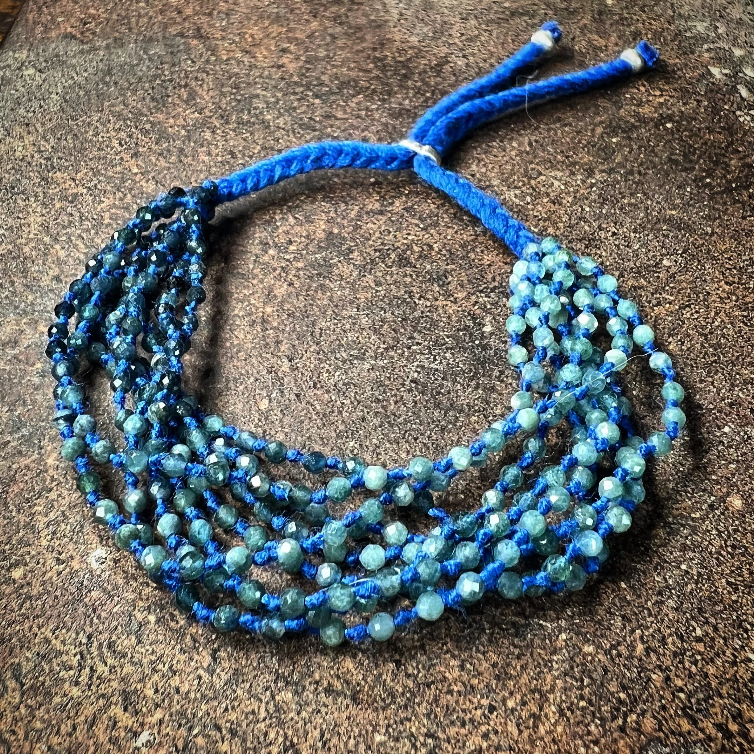 A Blue tourmaline bracelet with various shades of blue beads, woven into five strands, with an adjustable blue cord at the top, resting on a rough brown surface.