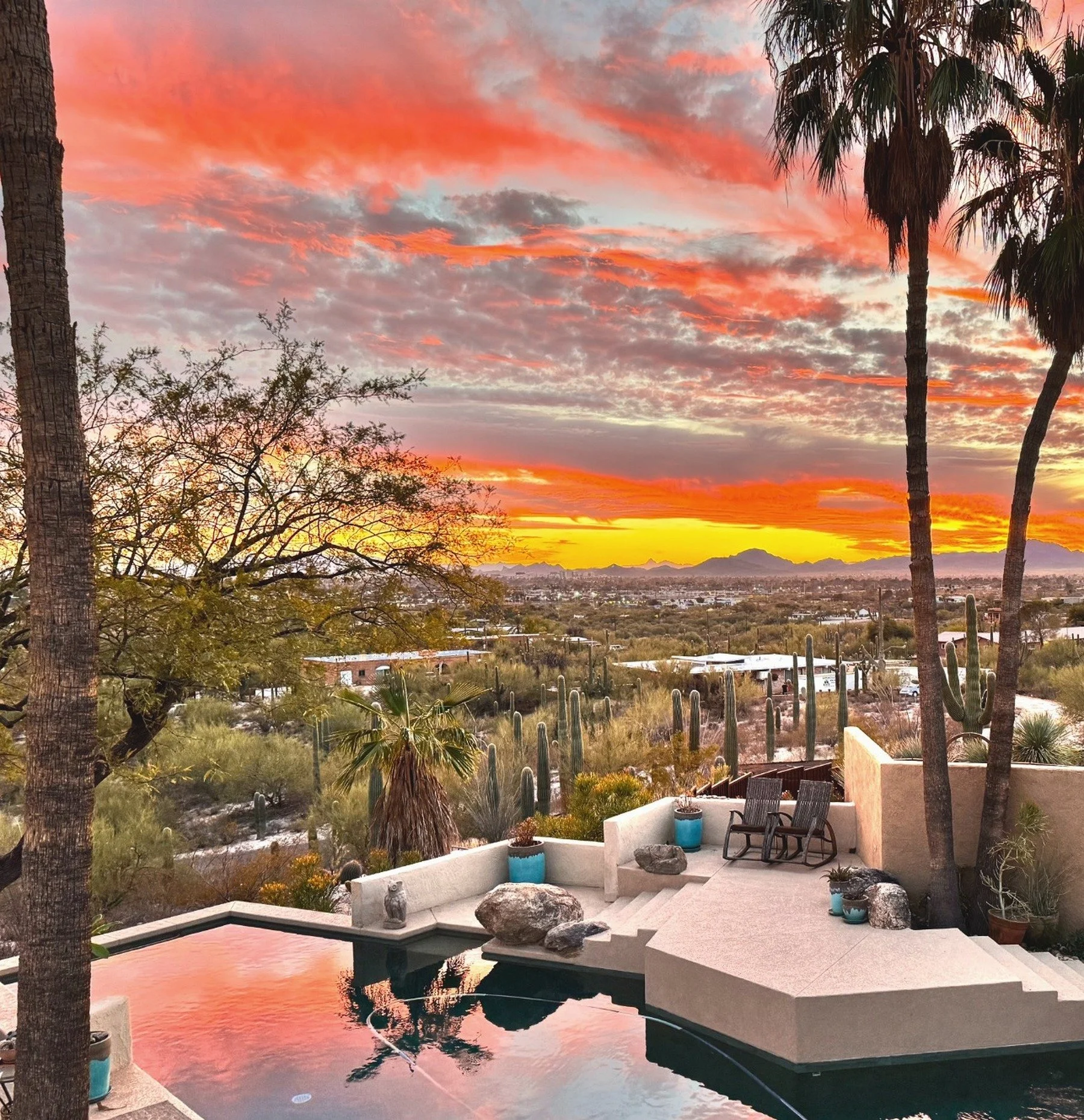 Scenic sunset over a desert landscape with cacti, palm trees, and residential buildings, viewed from a backyard with a pool and outdoor furniture.