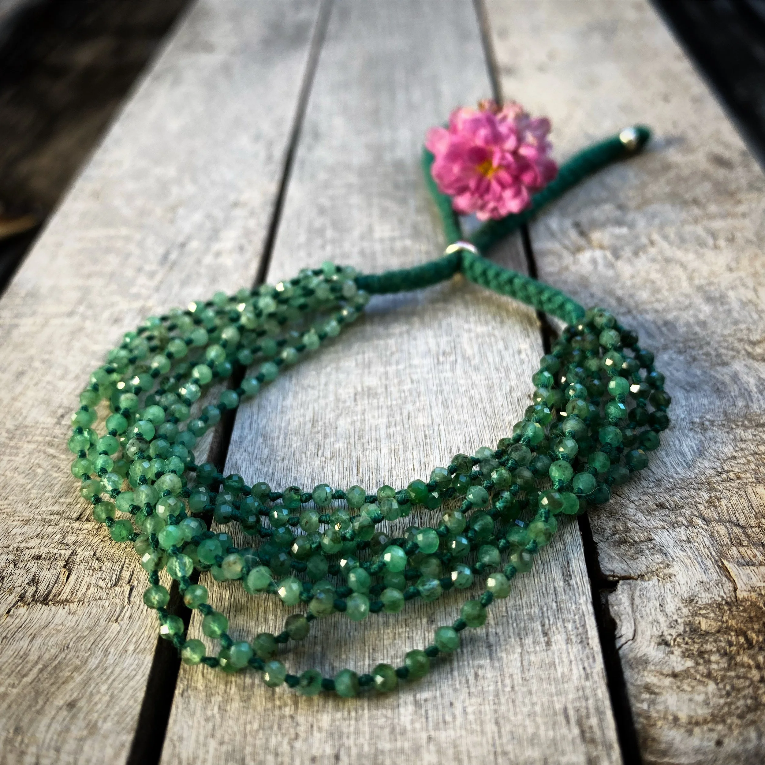 A beaded emerald bracelet with a pink flower attached, resting on a weathered wooden surface.