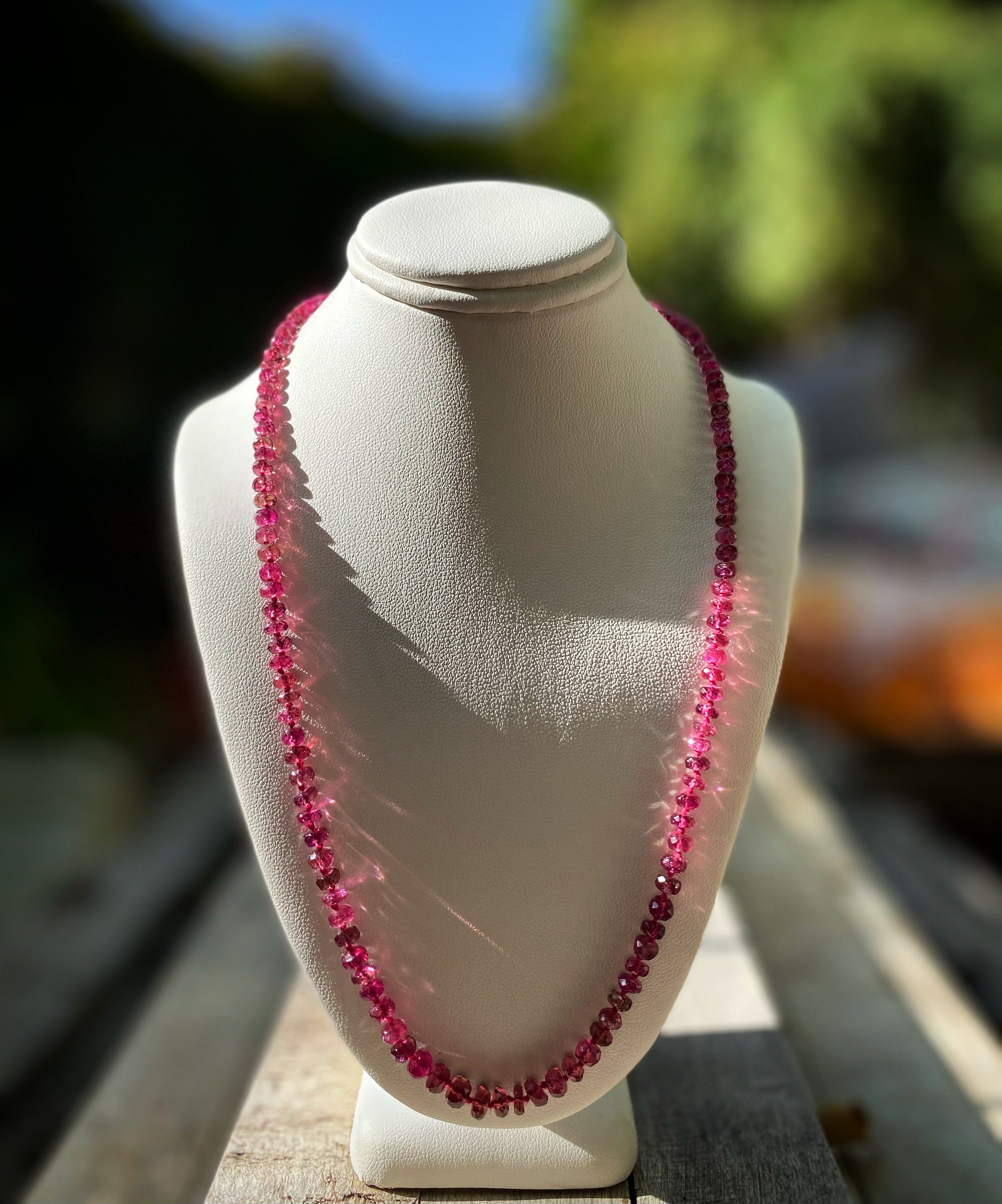 Pink tourmaline necklace displayed on a white bust outside with blurred trees and sky in the background.