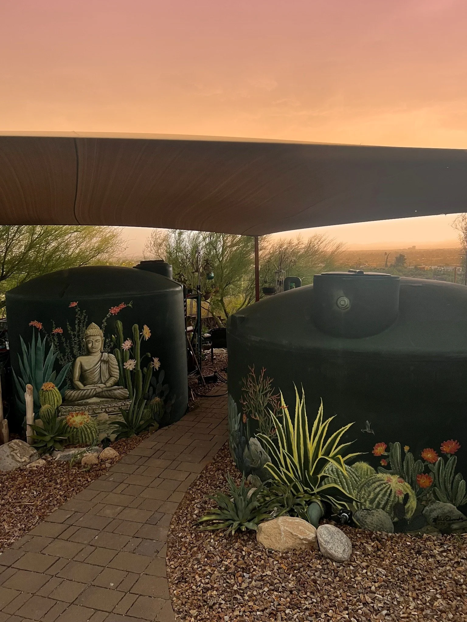 A sunset view over a terrace with two water tanks, one decorated with a mural of Buddha surrounded by plants and flowers, a pathway with pavers, decorative rocks, desert plants, and a distant cityscape under a pink-orange sky.