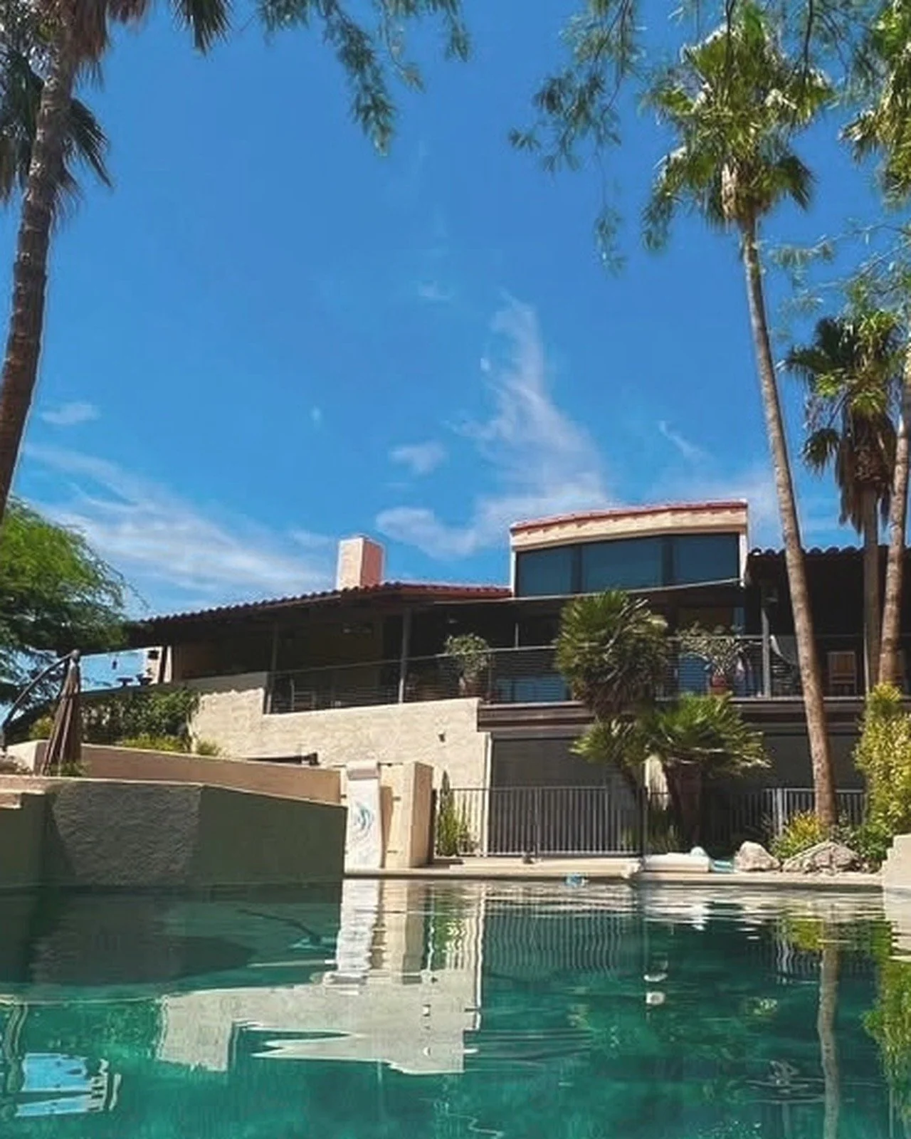 Modern house with large windows and balcony, surrounded by palm trees, with a swimming pool in the foreground and a clear blue sky above.