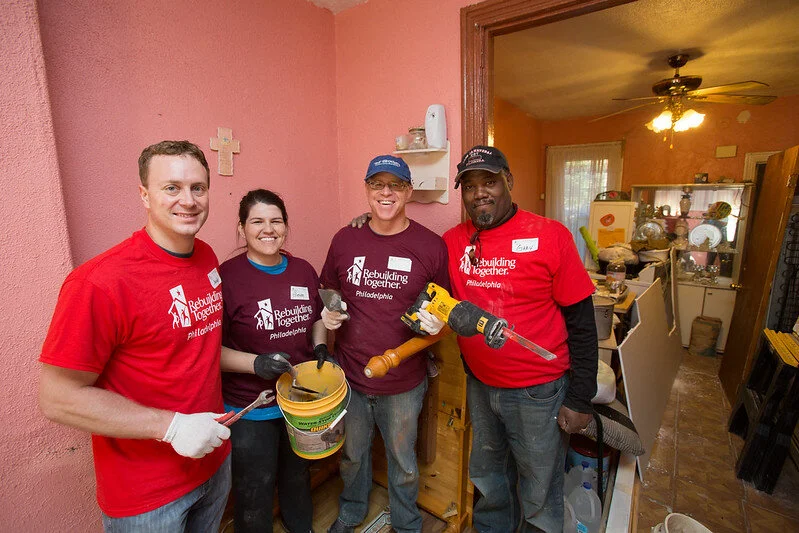 Four house captains stand outside a house entrance, smiling and holding construction supplies.
