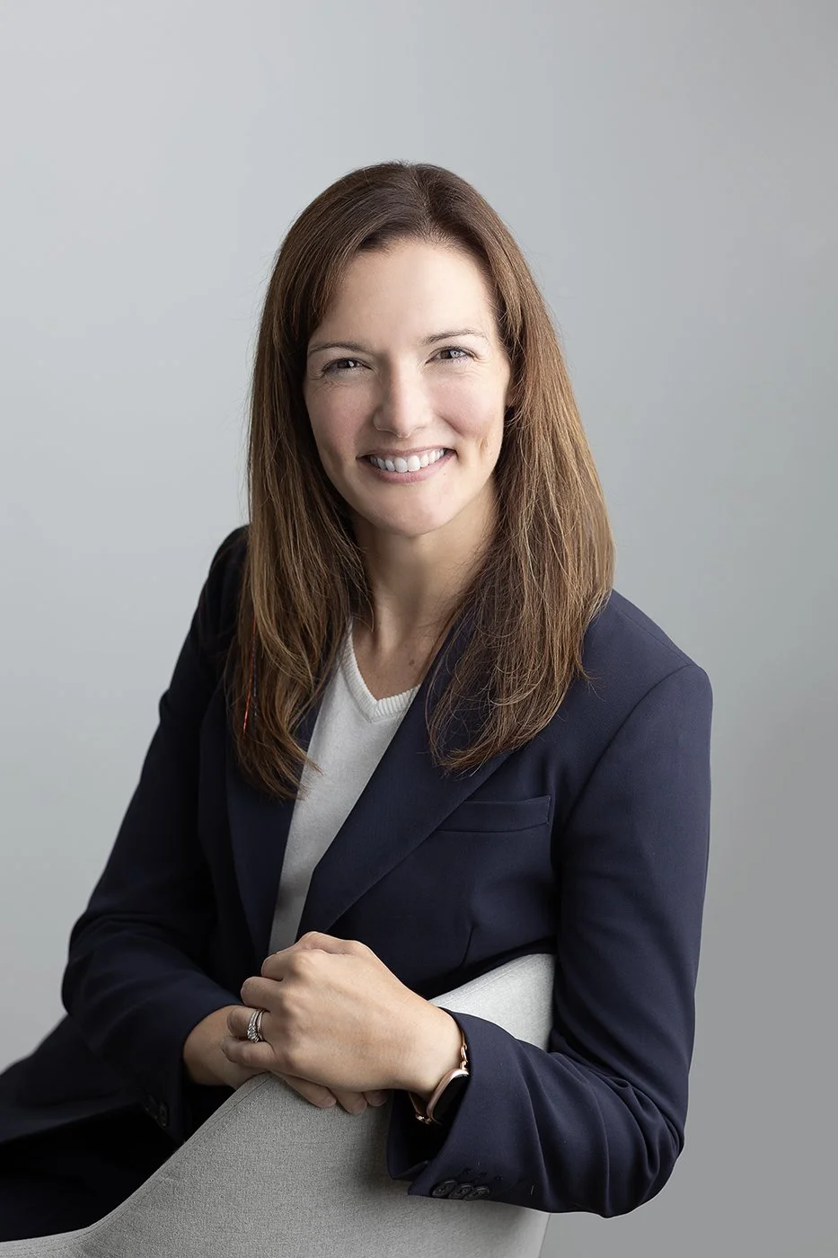 A woman smiles at the camera. She is wearing a dark blazer and a white shirt with a plain background behind her.