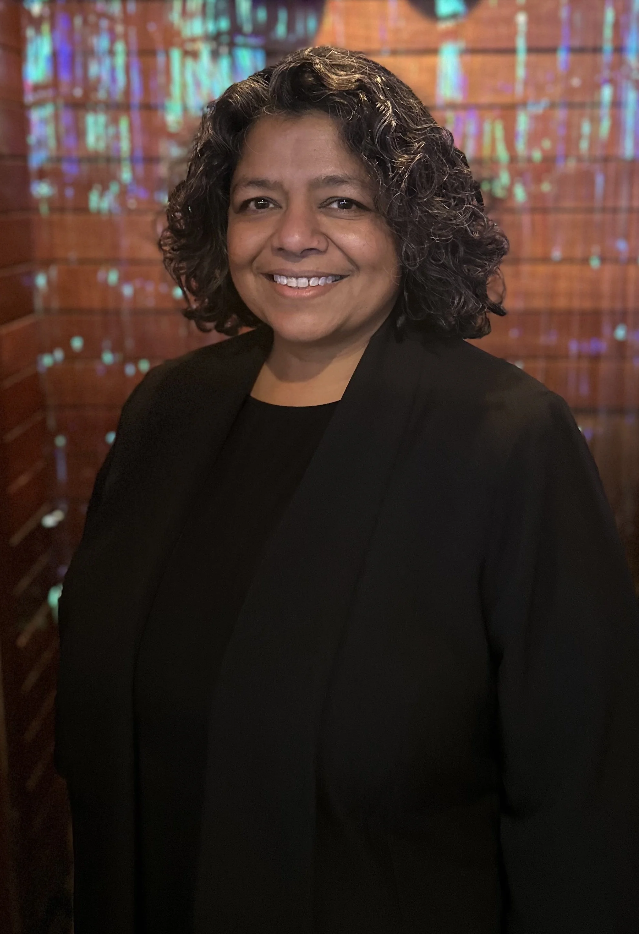 A woman in a black shirt and jacket smiles at the camera with a colorful shiny backdrop behind her