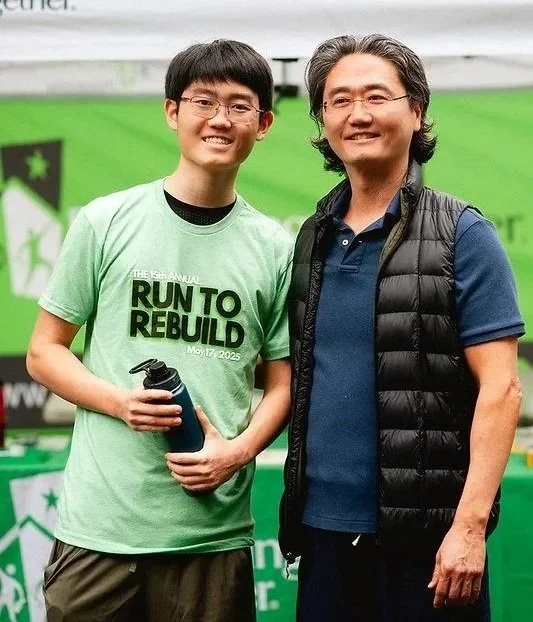 A father and son smile at the camera. The son is wearing a shirt that says Run to Rebuild.
