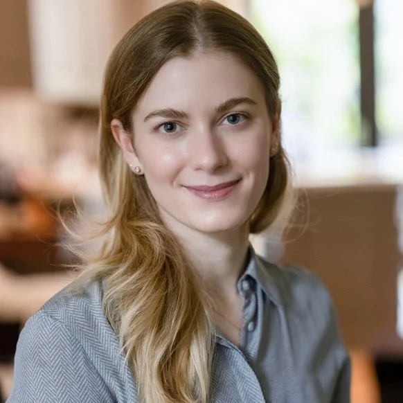 A white woman with blond hair smiles at the camera with her mouth closed. She is wearing a blue shirt and the background is blurred.