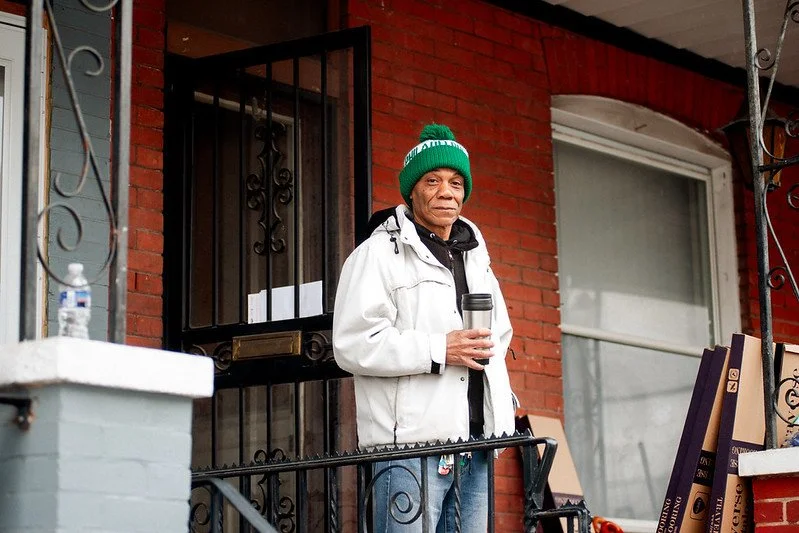 A homeowner wearing a green beanie and white jacket stands holding a coffee mug looks at the camera from the front porch of his rowhome.