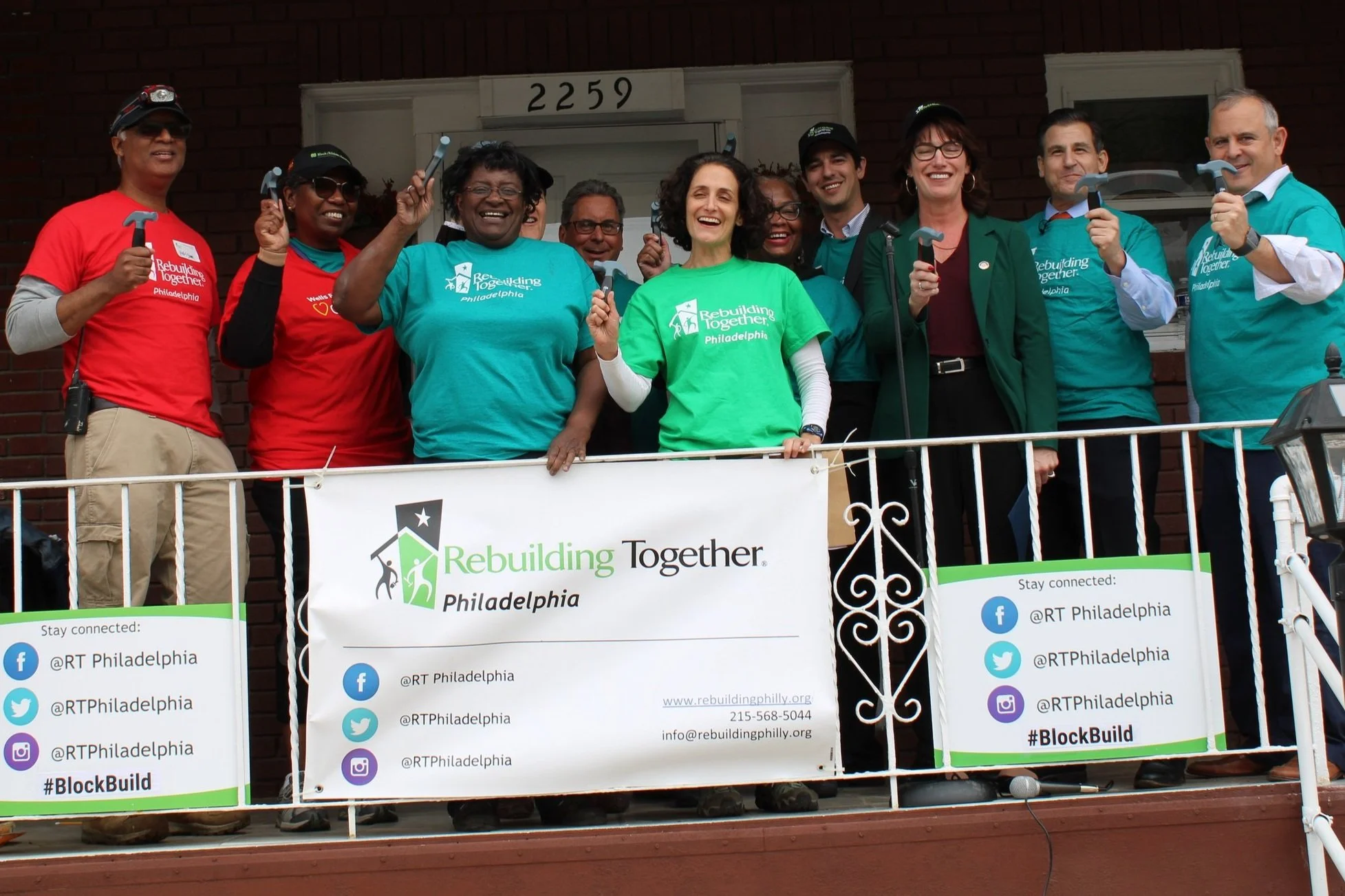 Group of smiling volunteers standing on a porch and holding up hammers.