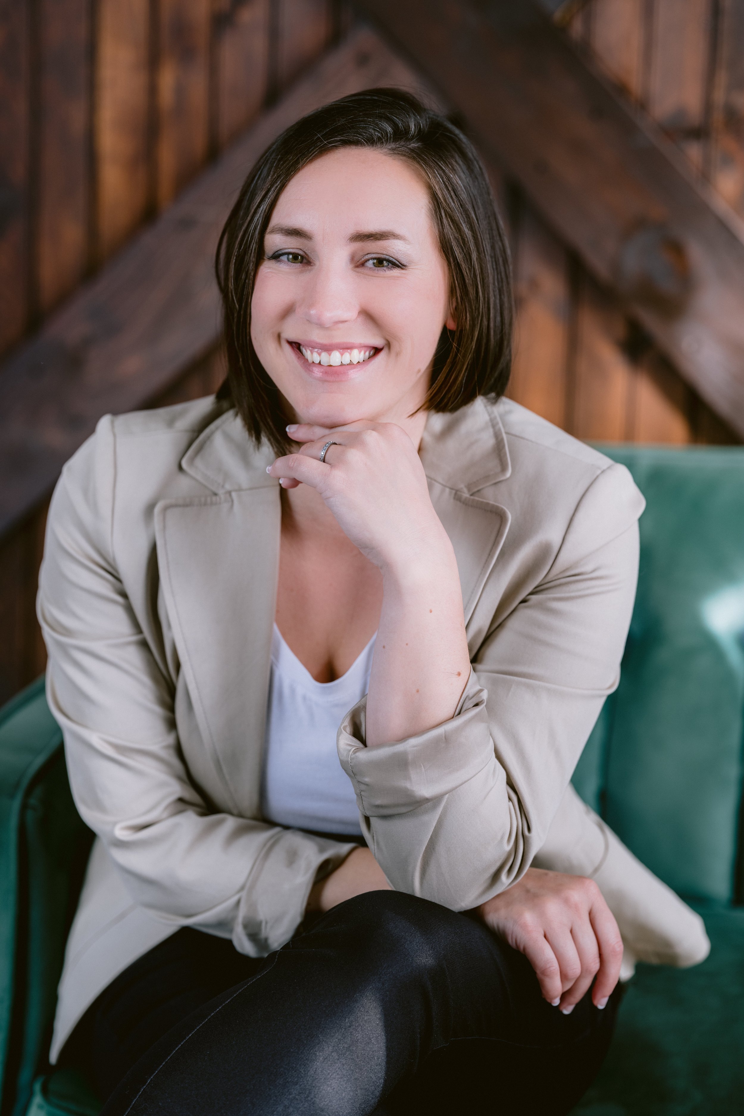 A woman smiles at the camera with her chin balanced on her hand. She has dark hair and beige blazer.