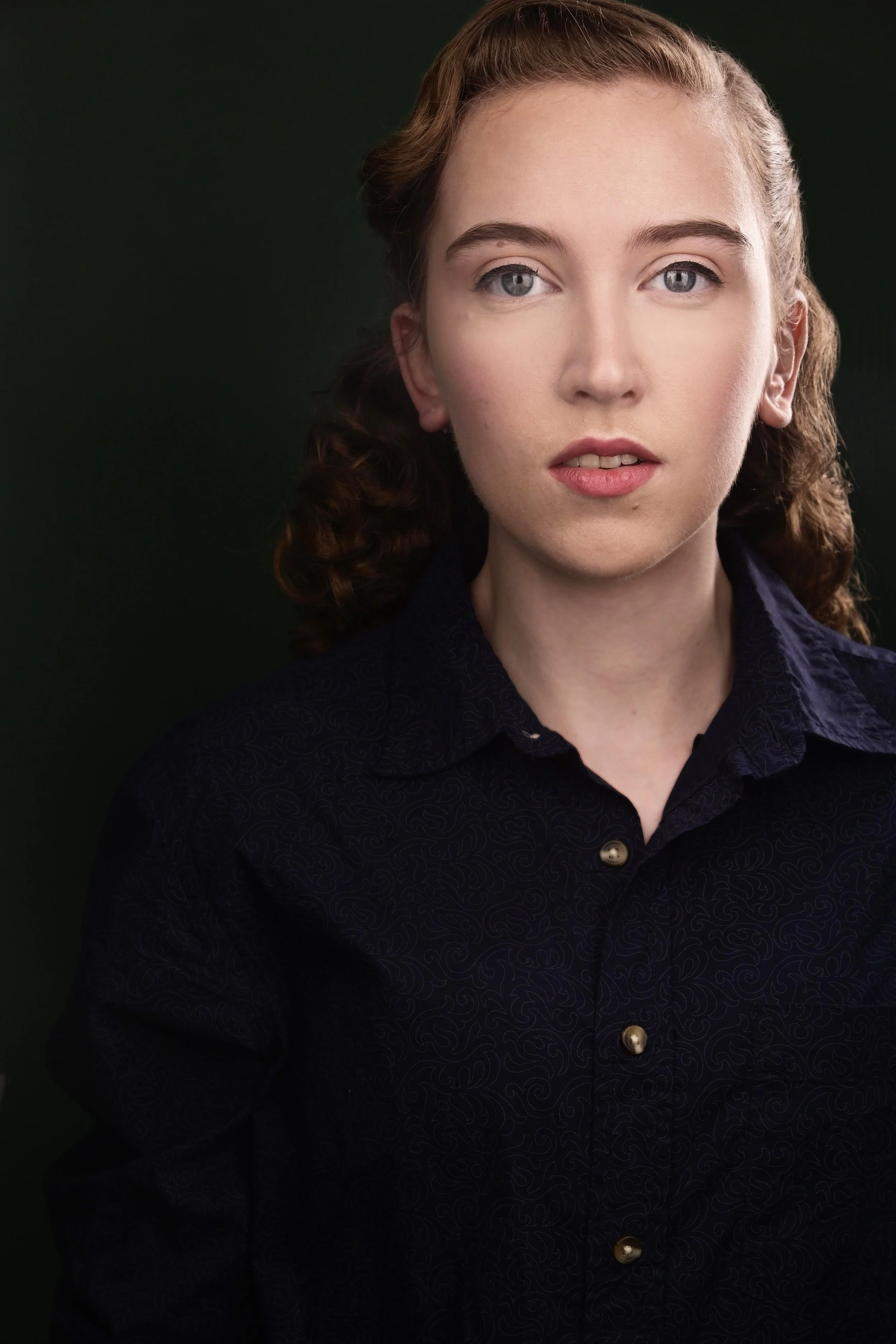 Headshot of Sydney - a young white person with light brown long curly hair wearing a dark navy button up and staring at the camera straight on