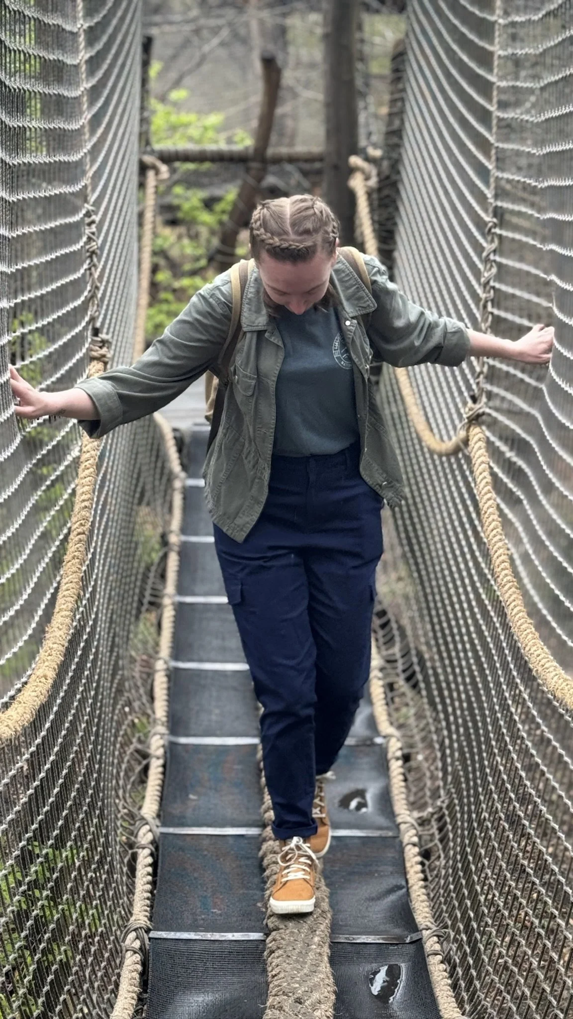 Image of Sydney, a young white person with their hair in complicated braids wearing navy pants, a dark teal shirt with a green cargo jacket and yellow sneakers, is focusing on walking across a rope bridge