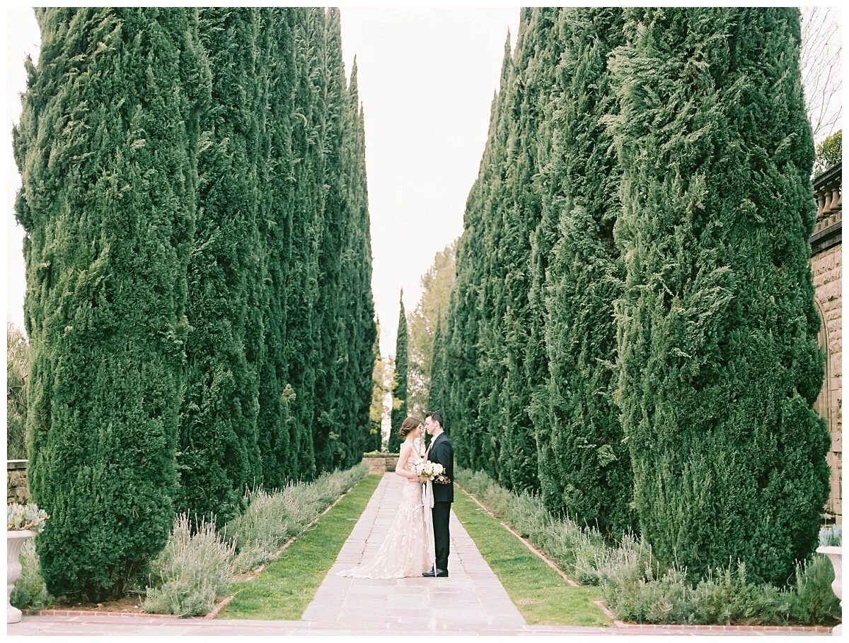 A bride and groom stand close together in a garden walking path, surrounded by tall green cypress trees on either side, with the bride holding a bouquet of flowers.