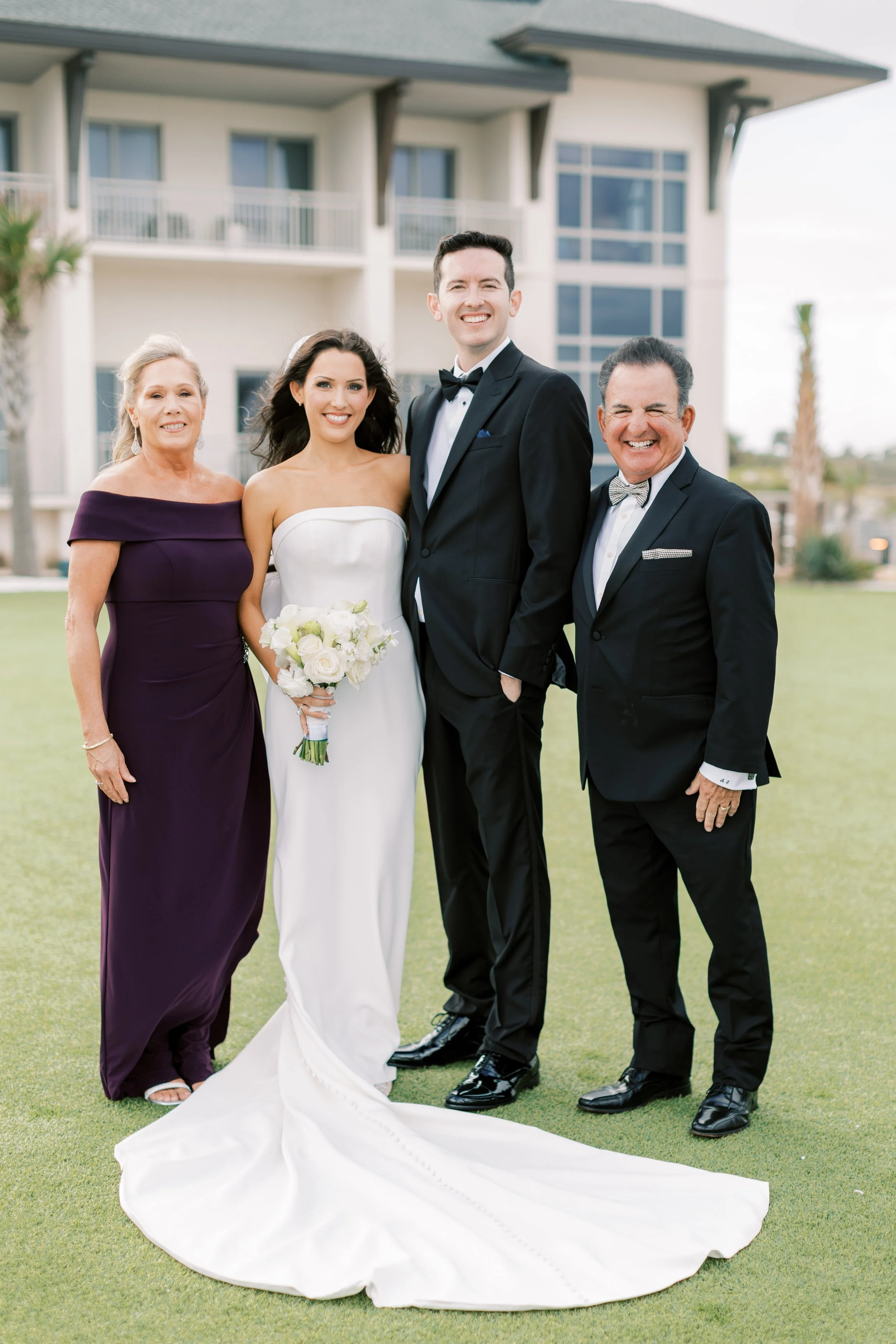 Group photo of four people outdoors in front of a modern house, including a bride in a strapless white wedding gown holding a bouquet, a groom in a black tuxedo, and two older individuals also dressed formally, smiling.