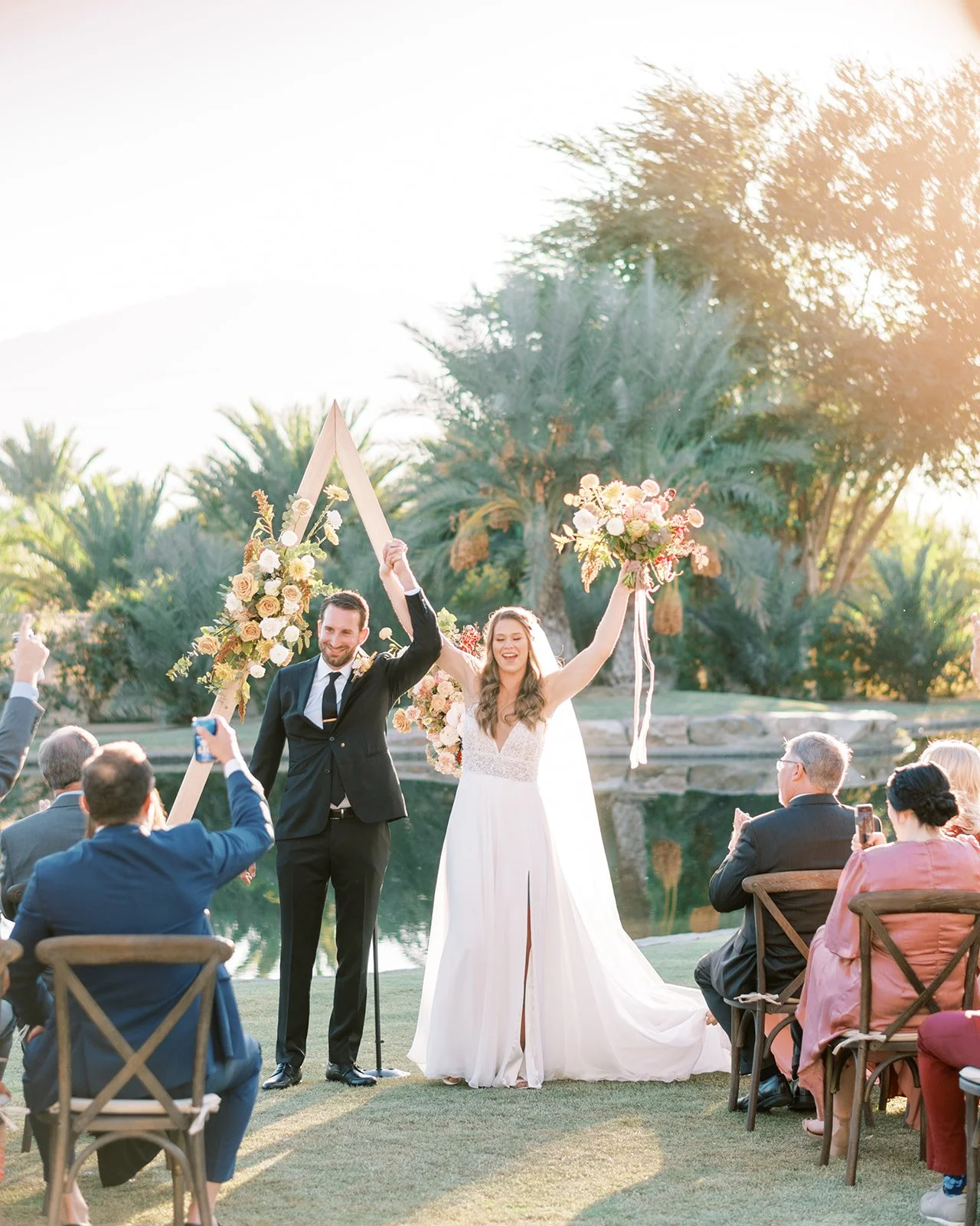 A bride and groom celebrate their wedding outdoors by holding up a triangular wooden sign decorated with flowers, with joyful expressions, surrounded by seated guests, in a garden setting with palm trees and a pond, during sunset.