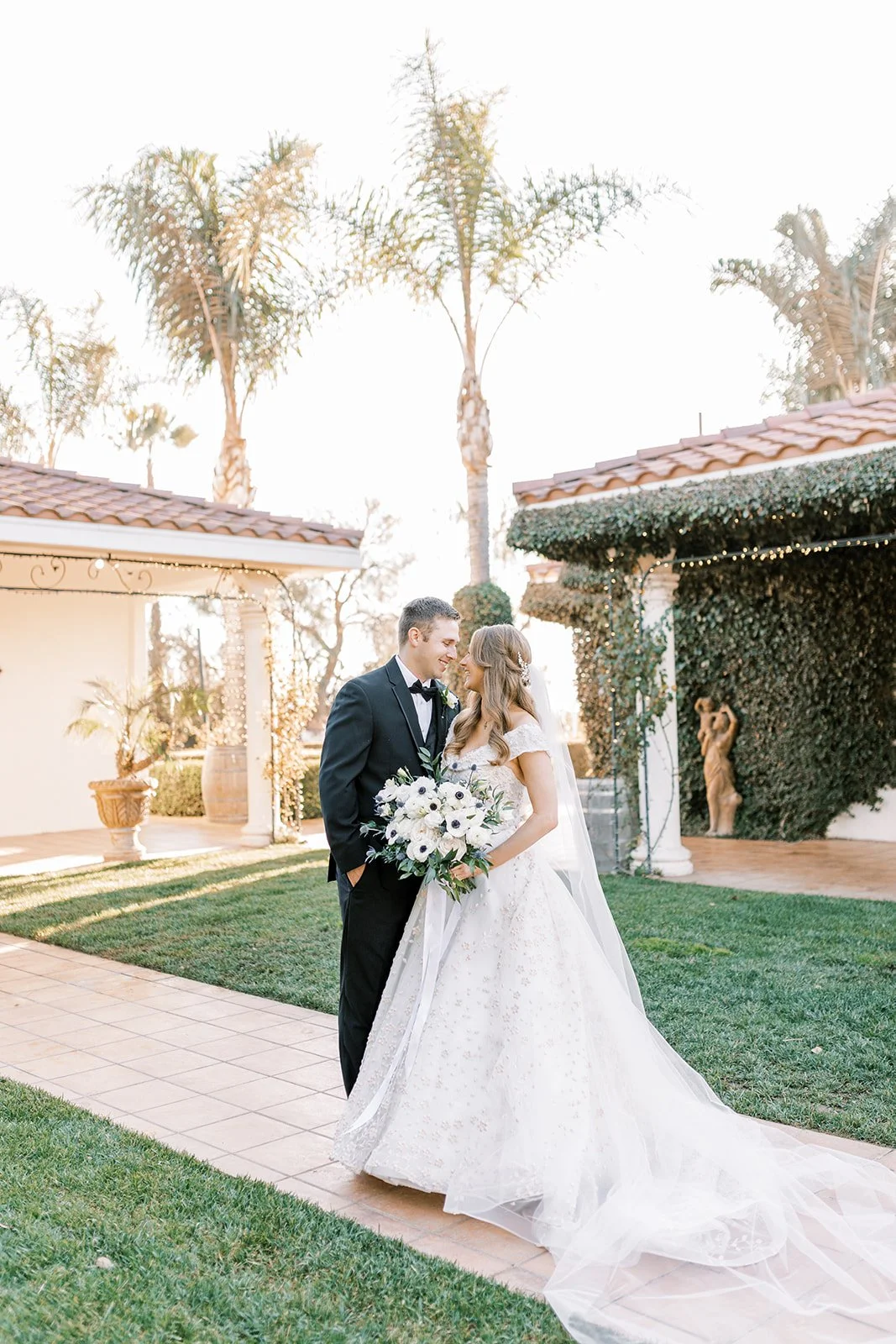 A bride and groom standing close together outdoors during their wedding, smiling at each other. The bride wears a white wedding gown and veil, holding a bouquet of white flowers. The groom is dressed in a black tuxedo with a bow tie. Palm trees and decorative greenery are visible in the background.