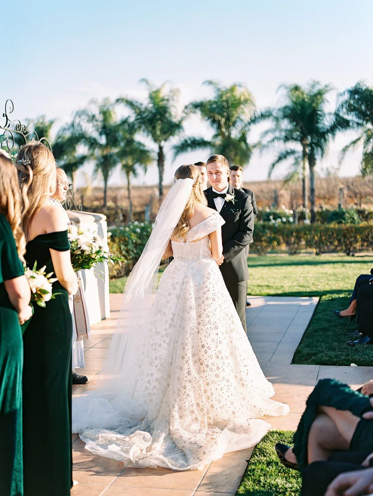 A wedding ceremony outdoors with the bride and groom standing together, surrounded by bridesmaids and guests, with palm trees in the background and bright sunlight.