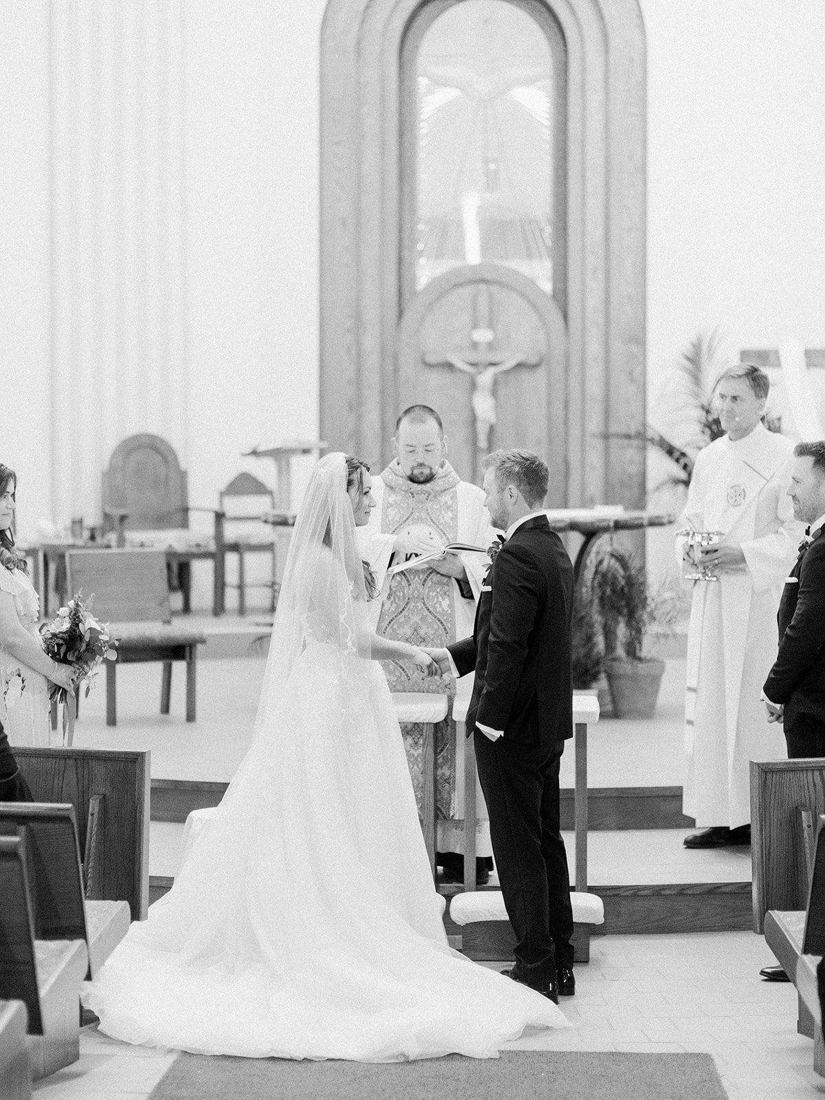 A wedding ceremony inside a church with a bride and groom holding hands, exchanging vows, officiated by a priest or minister. Bridesmaids and groomsmen stand nearby, with one attendee holding a bouquet. The church has religious symbols, including a crucifix and a stained glass window.