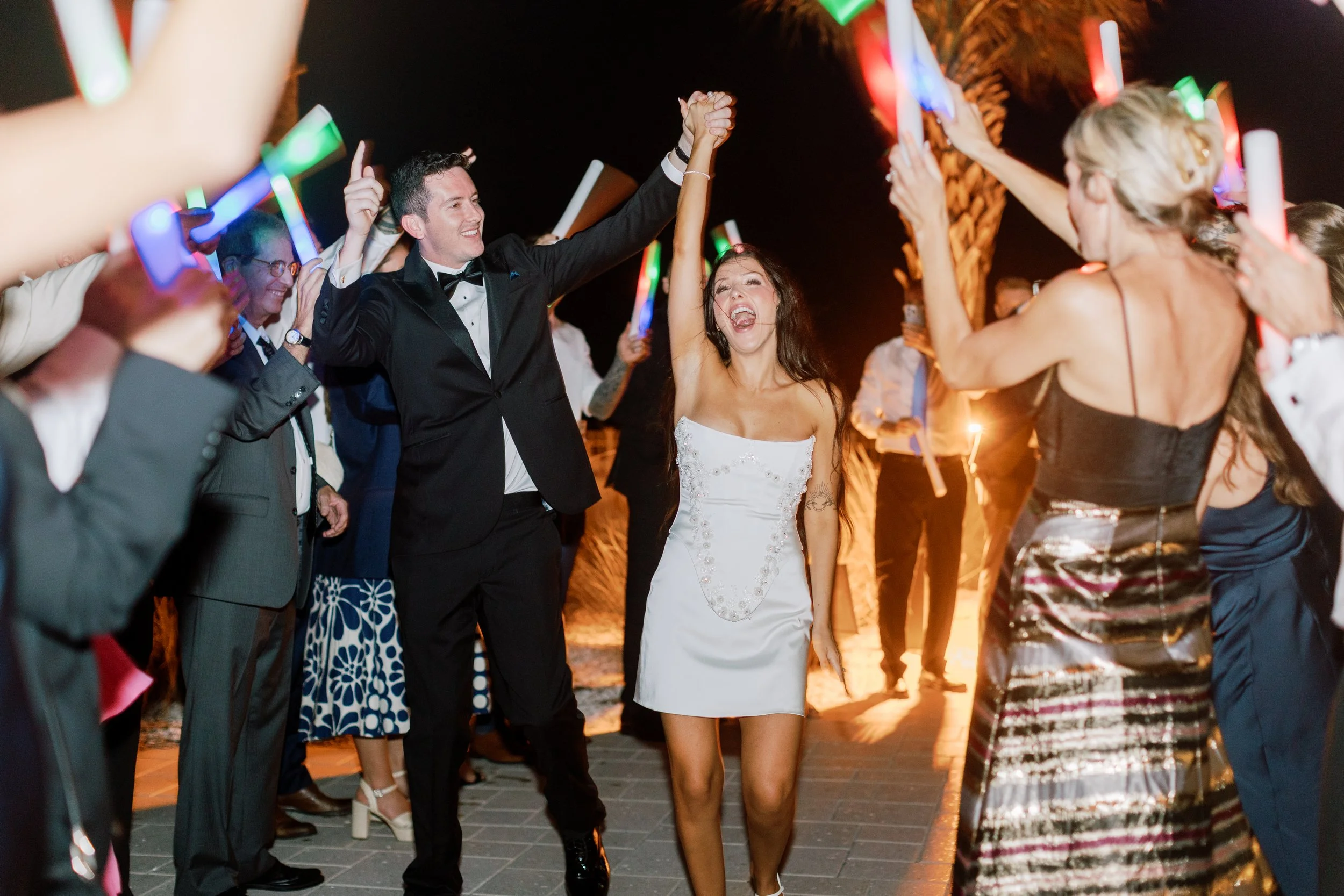 People dancing and celebrating at an outdoor event at night, with one woman in a white dress and a man in a tuxedo holding hands and raising their arms in joy.