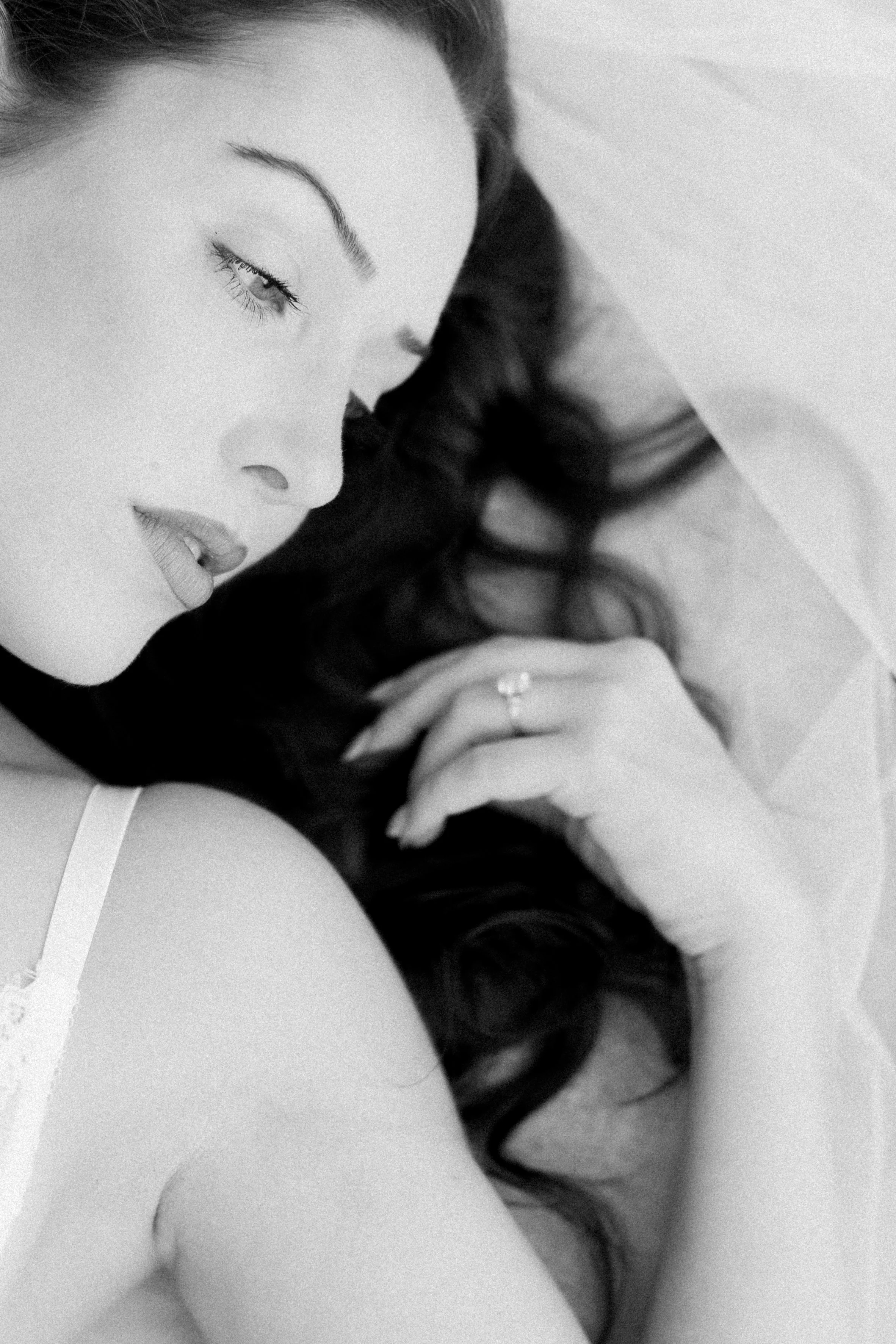 Close-up black and white photo of a woman with long curly hair, her eyes closed or about to close, lying on her side, wearing a white top, with a ring visible on her finger.