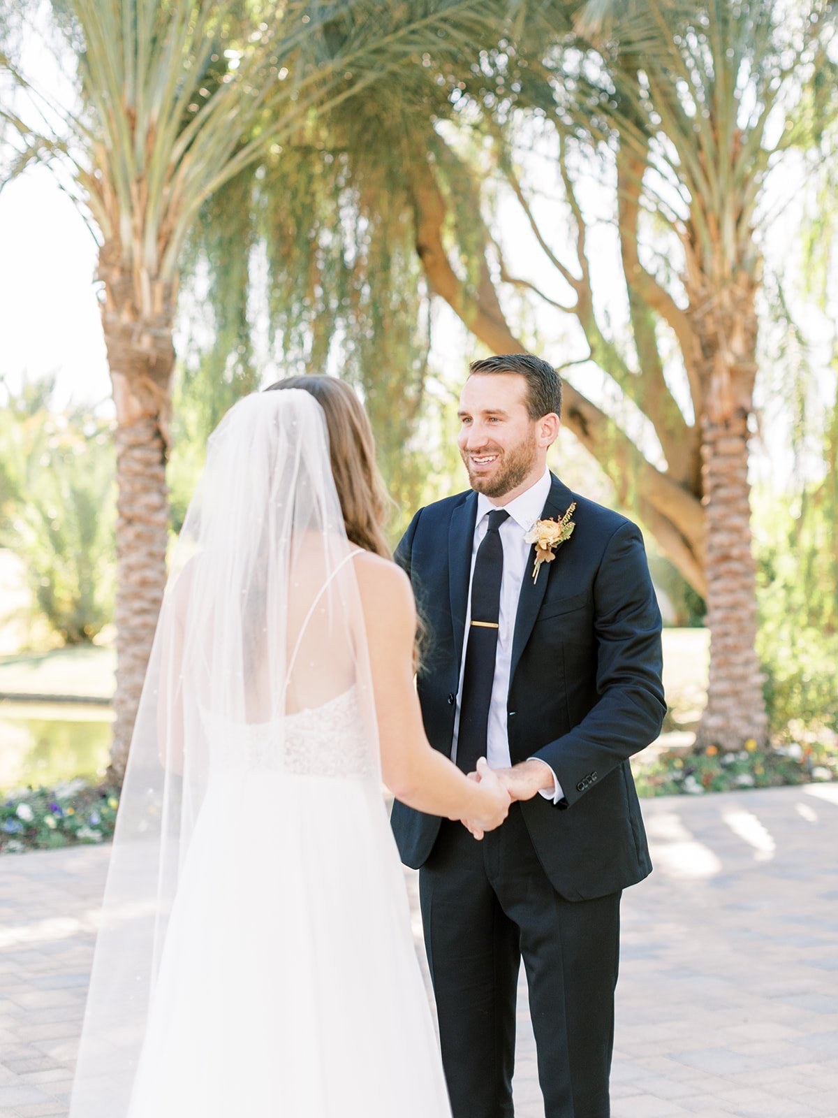 A bride and groom holding hands during their outdoor wedding ceremony, with palm trees and greenery in the background.