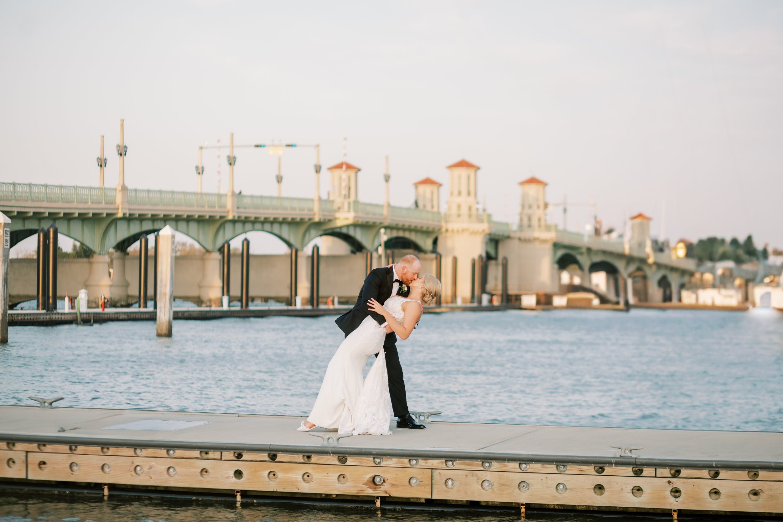 A bride and groom in wedding attire sharing a kiss on a dock by the water with a bridge in the background.