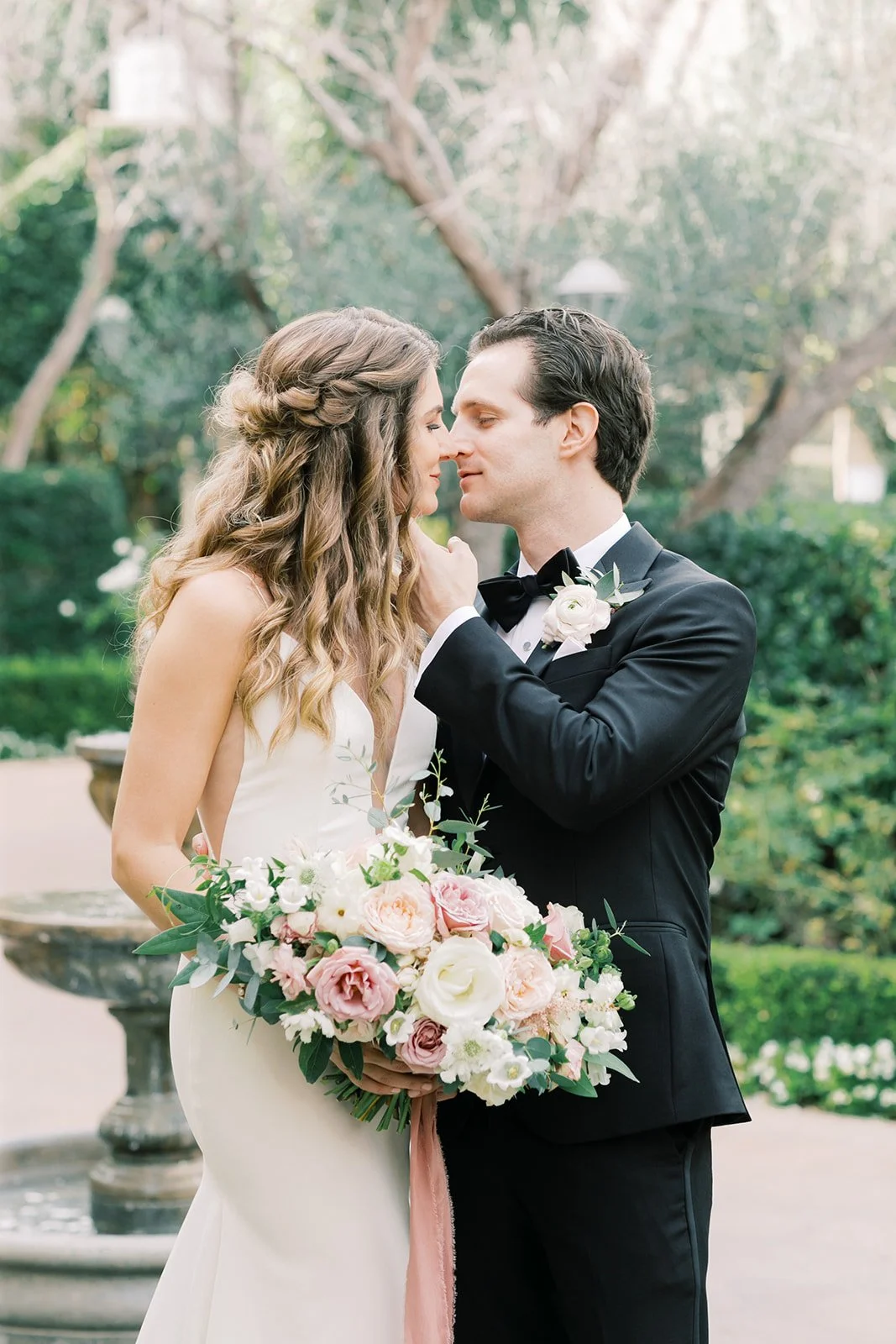 A bride and groom in wedding attire holding a bouquet of pink and white flowers, standing close and touching foreheads in an outdoor garden setting.