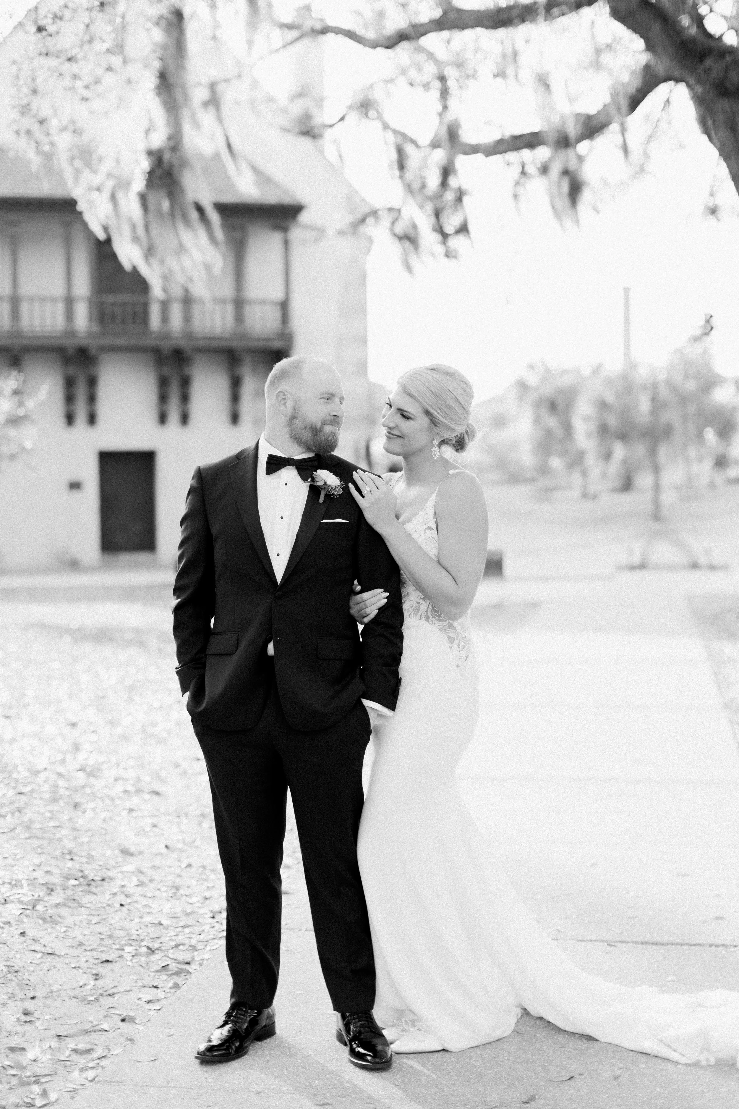 Black and white wedding photo of a bride and groom standing outdoors, smiling at each other, with trees and a building in the background.