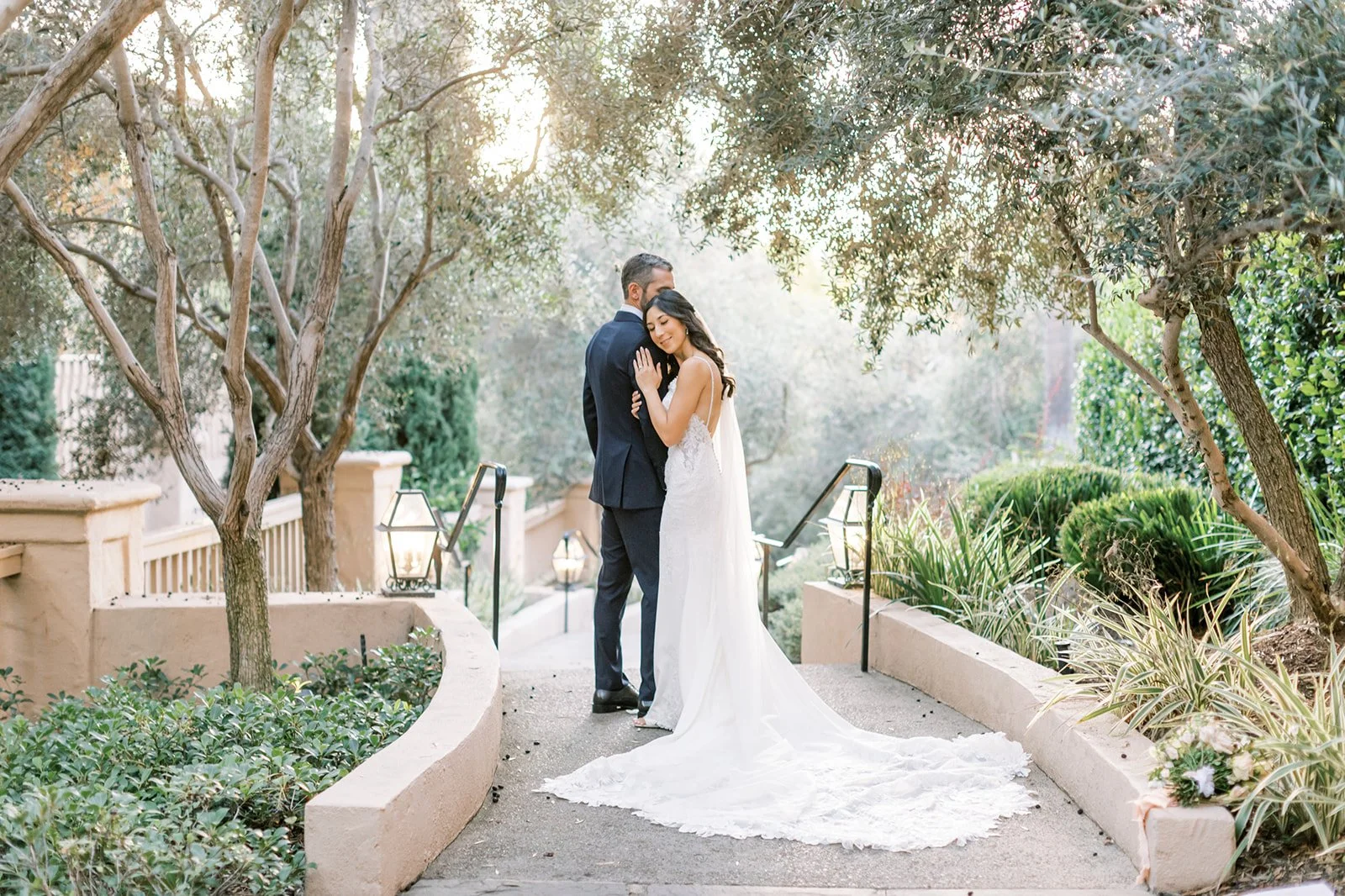 A bride and groom sharing a kiss on a garden pathway lined with small trees and plants during sunset.