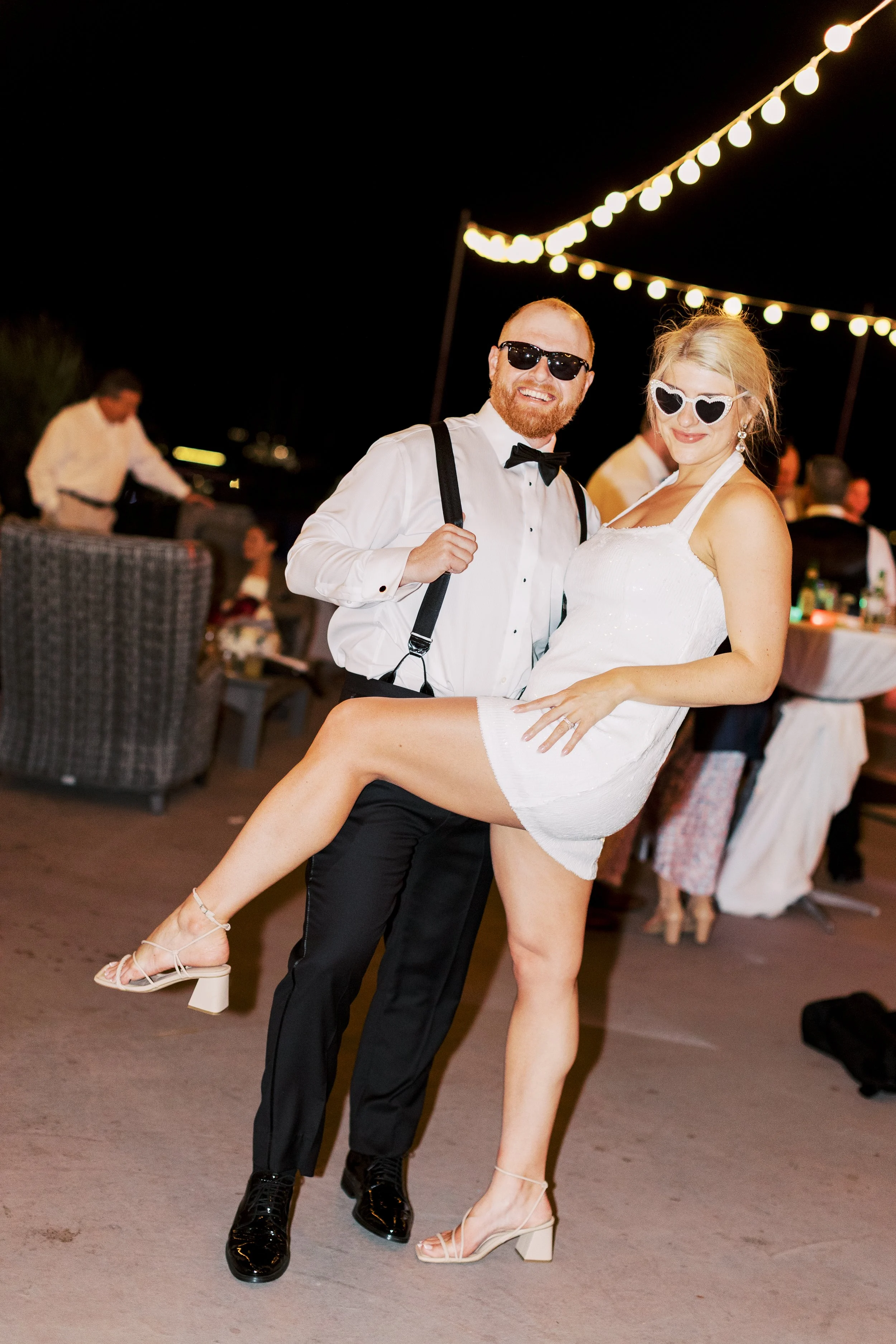 A happy couple dancing at an outdoor nighttime wedding reception, with string lights overhead. The man is wearing a white shirt, black suspenders, a bow tie, and sunglasses. The woman is wearing a white dress, heart-shaped sunglasses, and is being held up by the man while smiling.