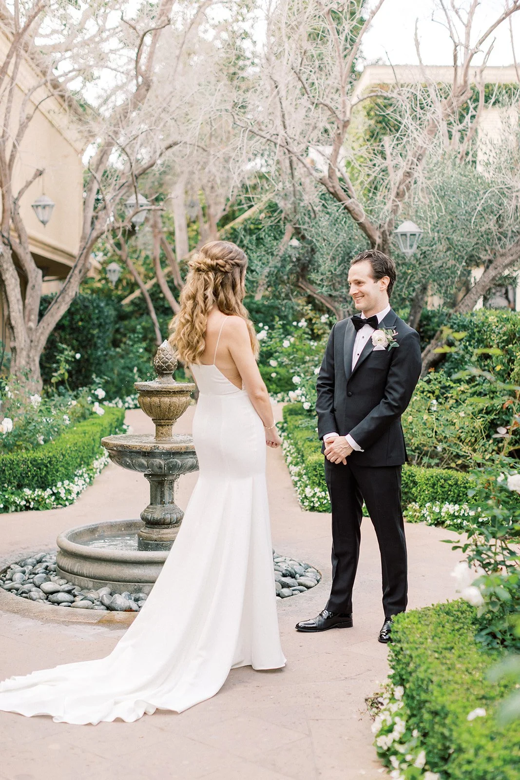 A bride and groom stand facing each other outdoors, surrounded by greenery and trees, during their wedding ceremony. The bride wears a white dress with a long train, and the groom is in a black tuxedo with a bow tie. A fountain is visible in the background.