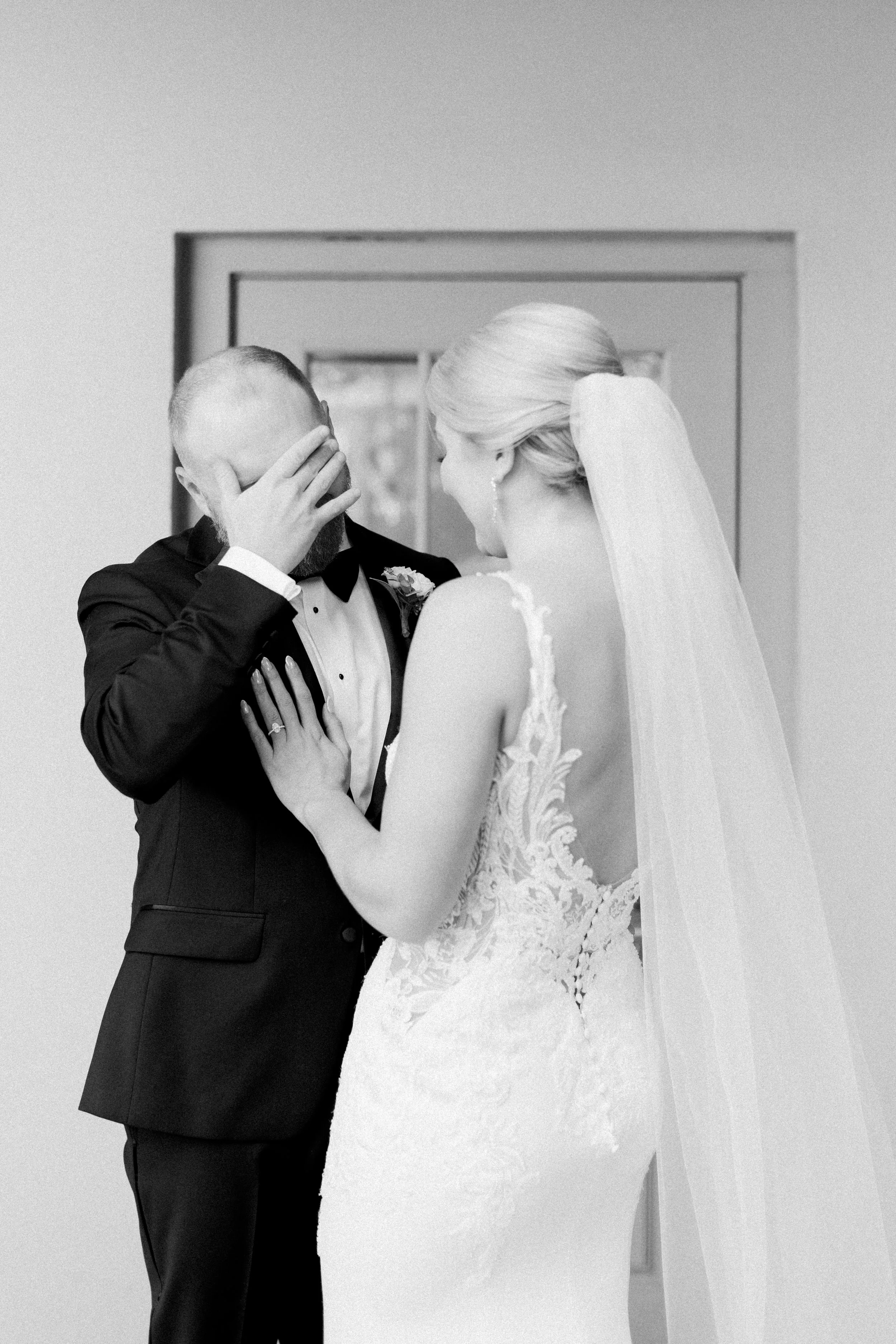 A bride and groom share an emotional moment with the groom wiping a tear from his face as the bride touches his chest, standing in front of a mirror.